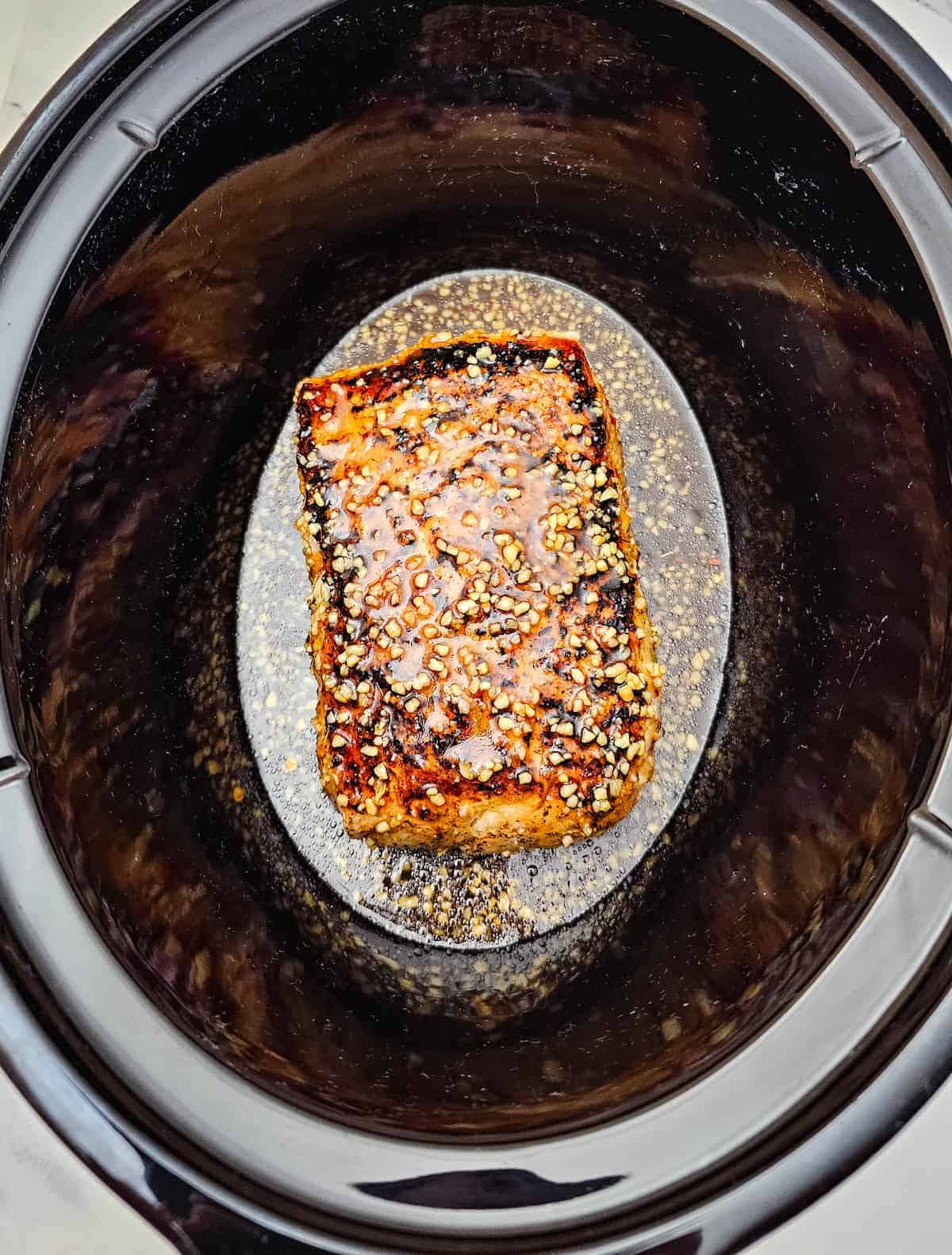 A cooked, seasoned piece of pork sits in the center of a slow cooker with visible juices and spices around it.