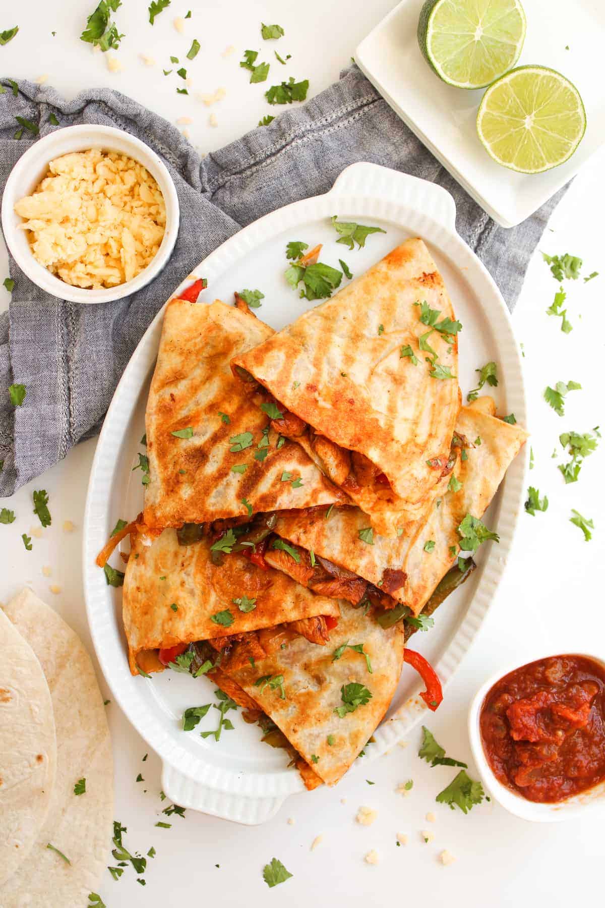 A platter of quesadillas garnished with chopped cilantro, served with salsa, shredded cheese, lime halves, and tortillas on the side.