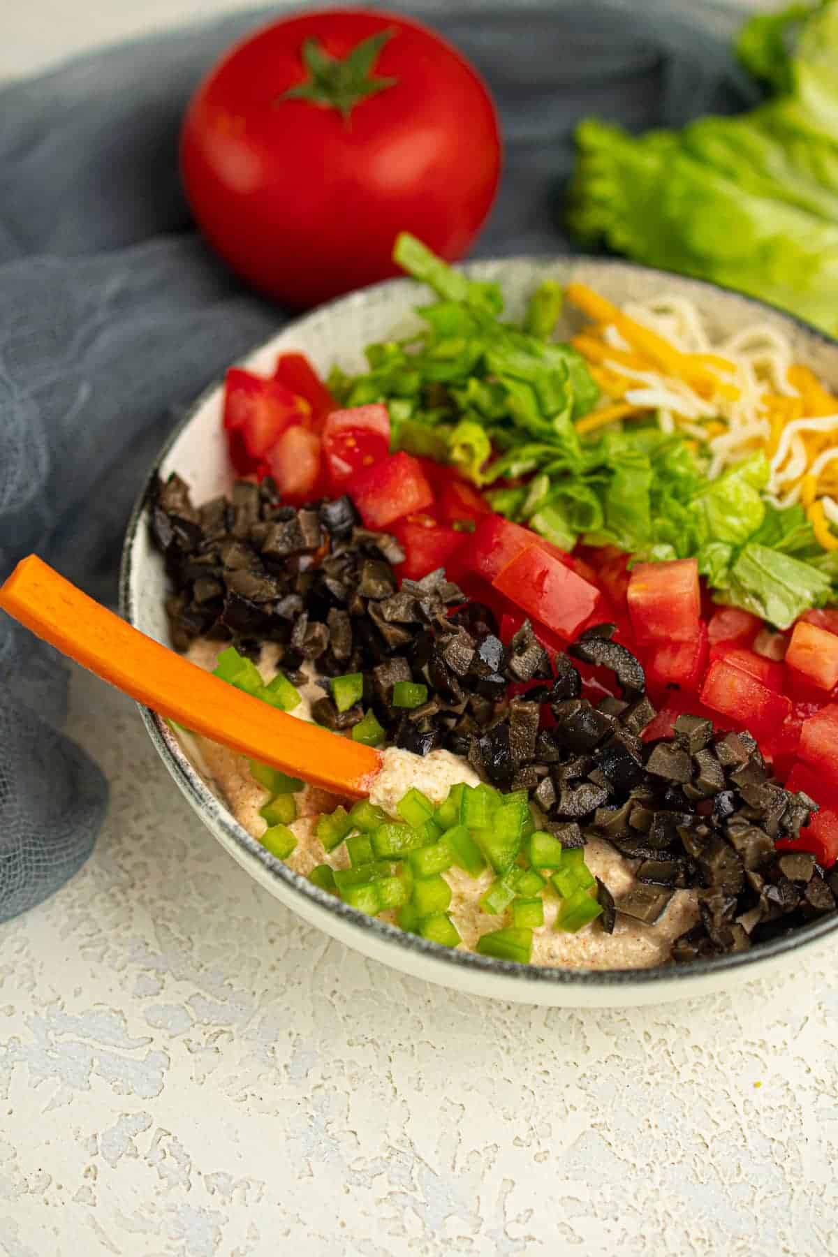A bowl of dip topped with chopped tomatoes, black olives, green bell peppers, lettuce, and shredded cheese, with a carrot stick on the side. A tomato and lettuce are in the background.
