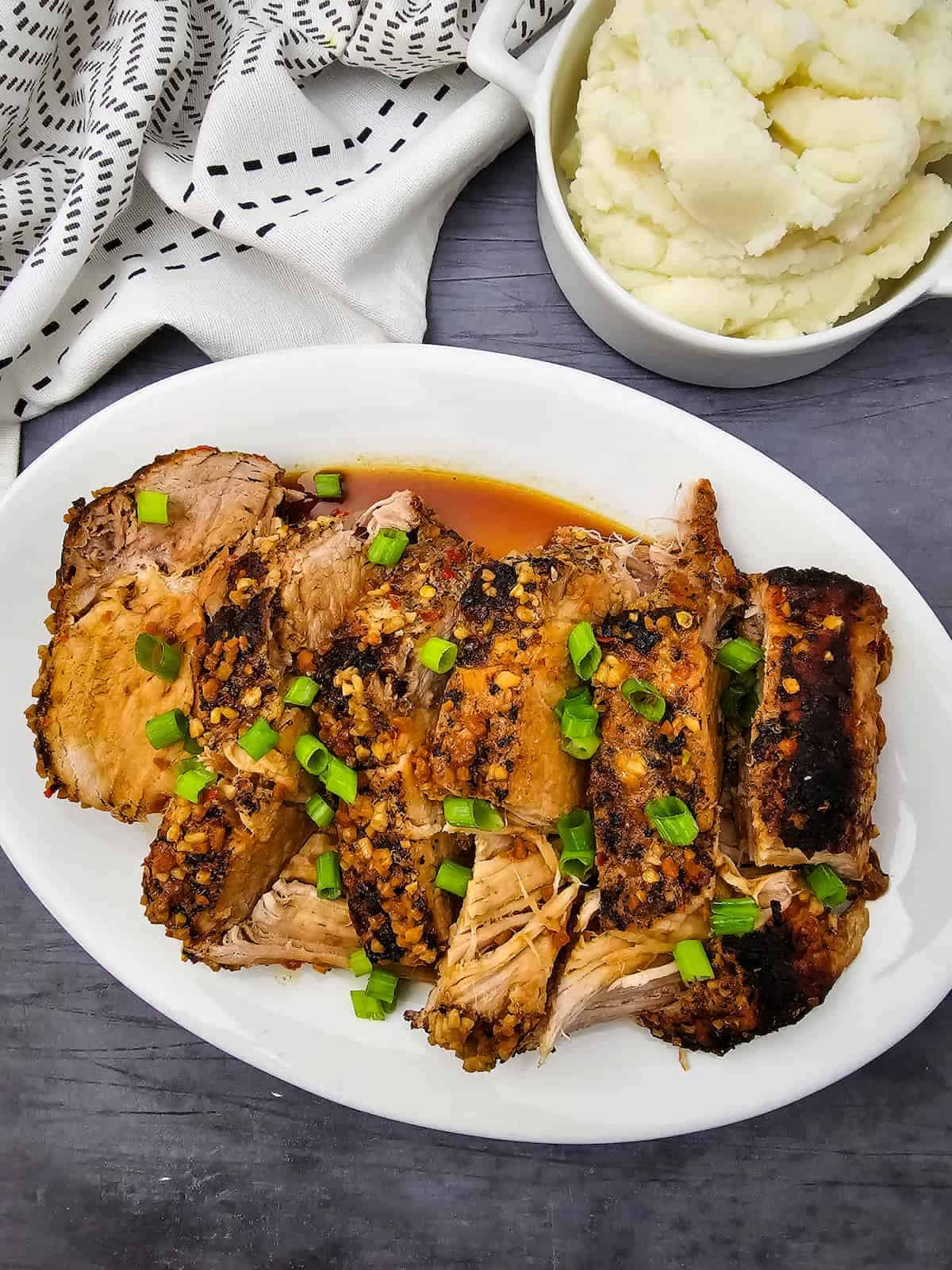 Sliced roast pork garnished with chopped green onions on a white plate, served with a side of mashed potatoes.