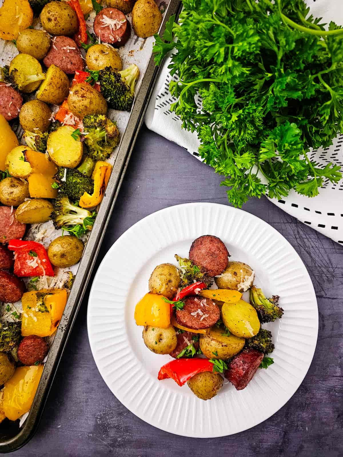 A plate of roasted potatoes, sausage, broccoli, and bell peppers is served next to a sheet pan with the same food. Fresh parsley is placed nearby on a dark surface.