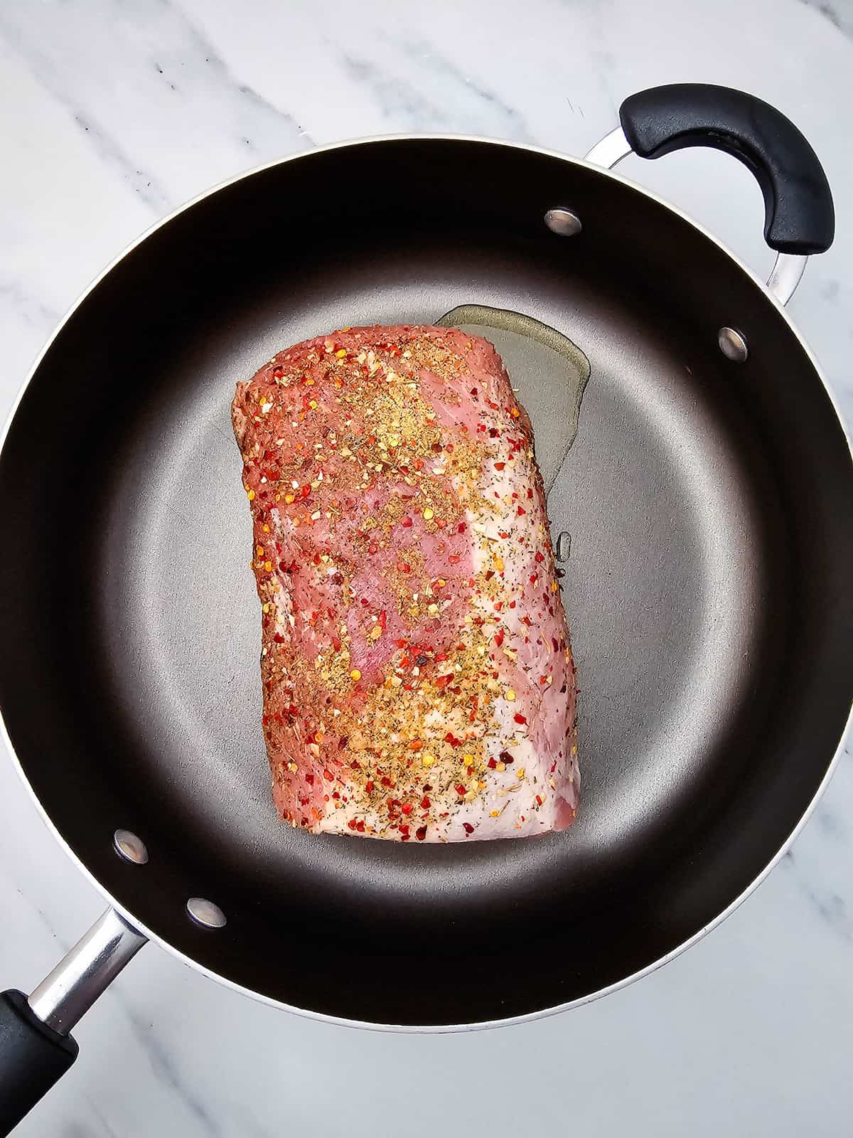 A raw, seasoned pork roast sits in a large black skillet with a small amount of oil on a white marble surface.