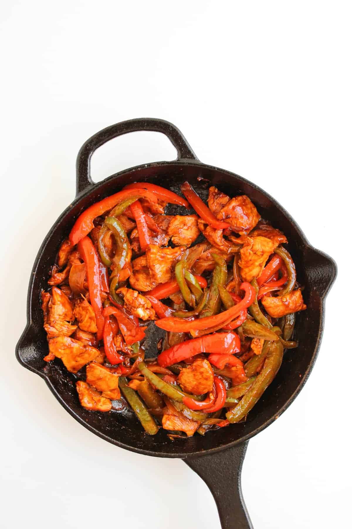 A cast iron skillet filled with cooked chicken strips, red and green bell peppers on a white background.