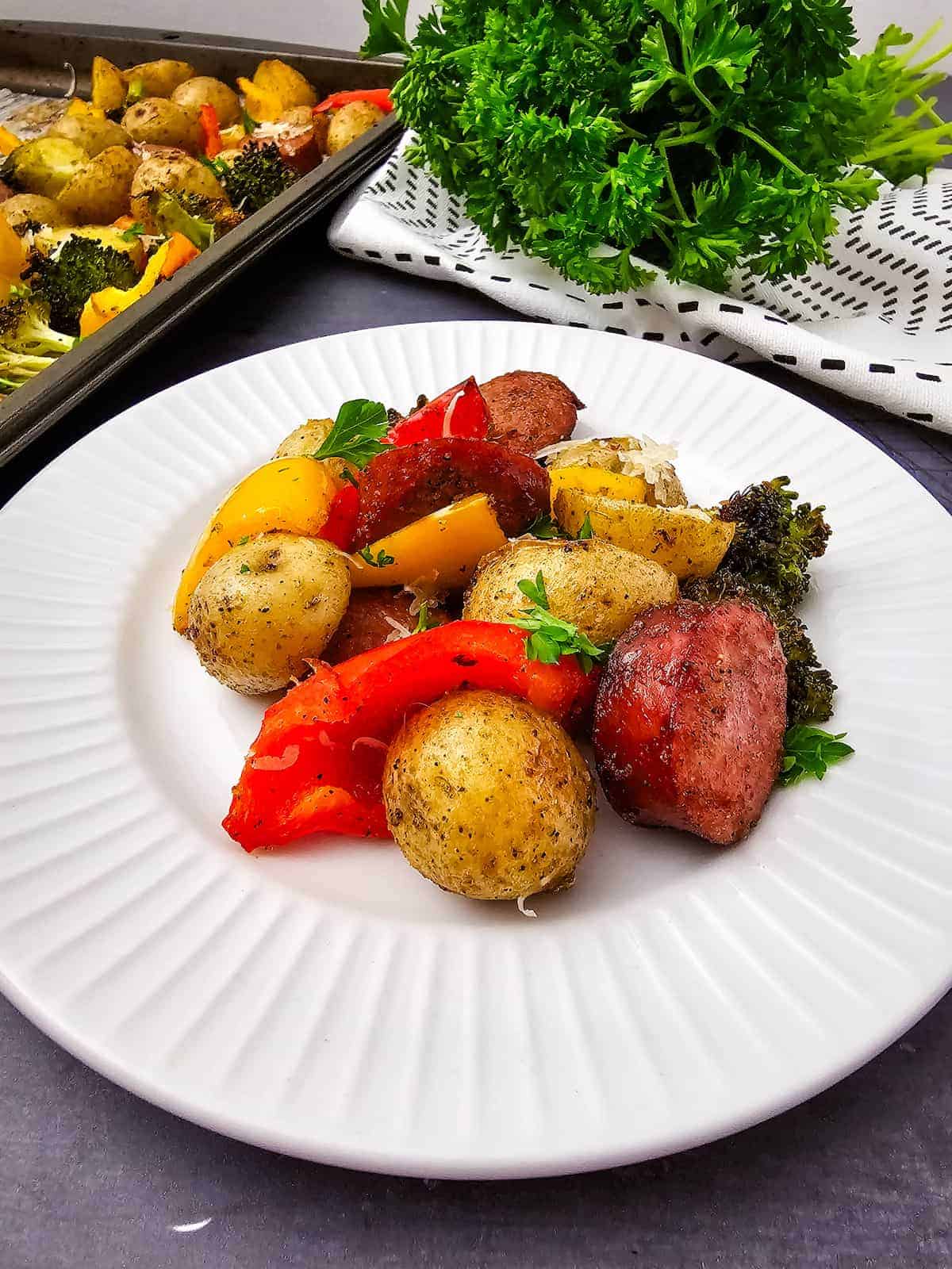 A white plate with roasted potatoes, sausage slices, red and yellow bell peppers, and broccoli garnished with parsley. A baking tray with more vegetables and parsley is in the background.