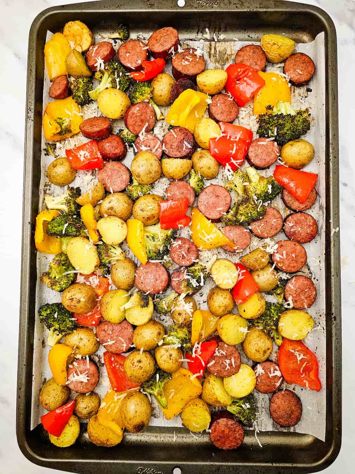 Sheet pan with roasted baby potatoes, sliced sausage, broccoli, and red and yellow bell peppers, sprinkled with shredded cheese.
