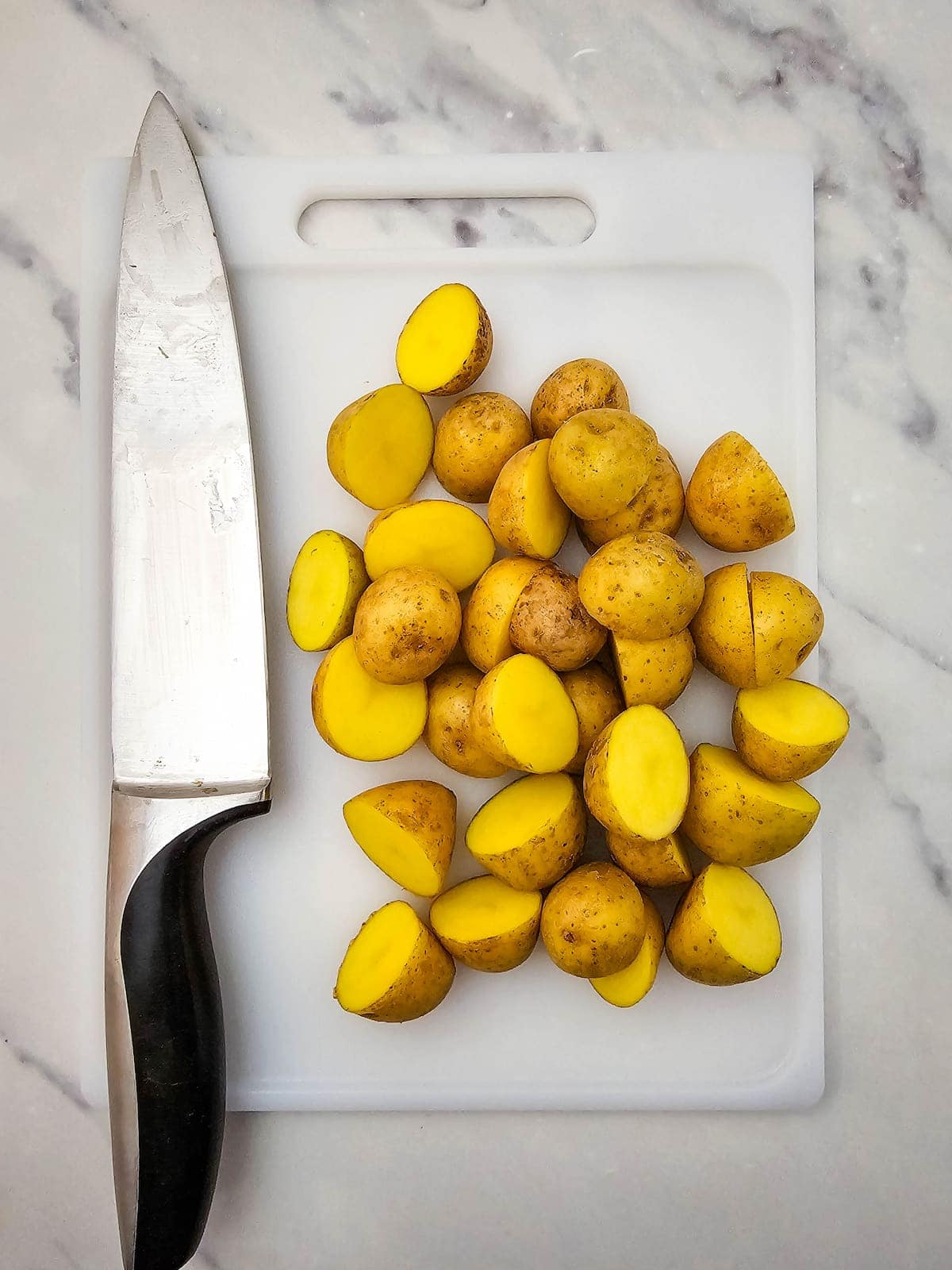 A kitchen knife and several halved yellow potatoes are arranged on a white cutting board atop a marble surface.