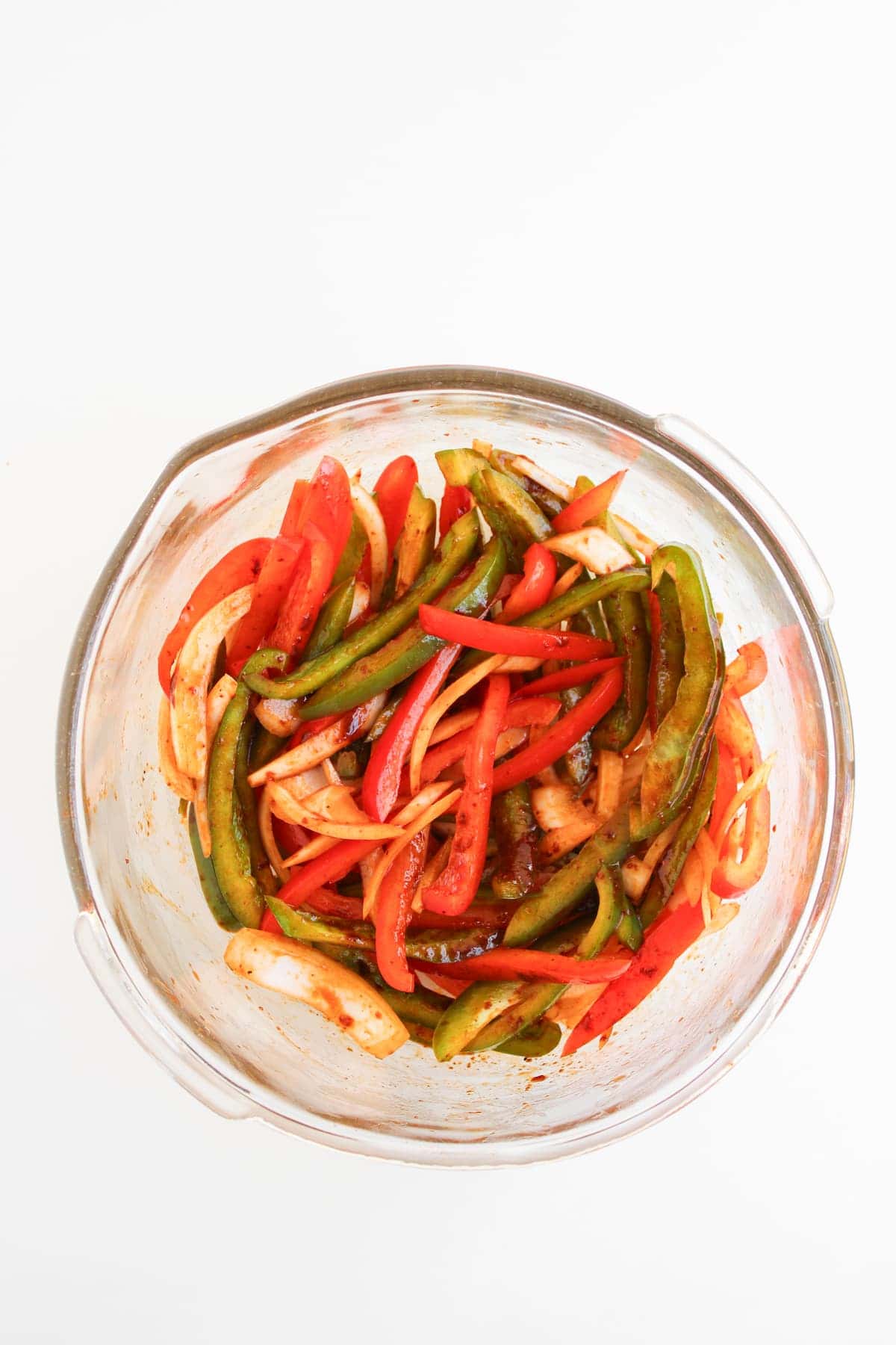 A glass bowl filled with sliced red and green bell peppers and onions, seasoned and mixed together, on a white background.