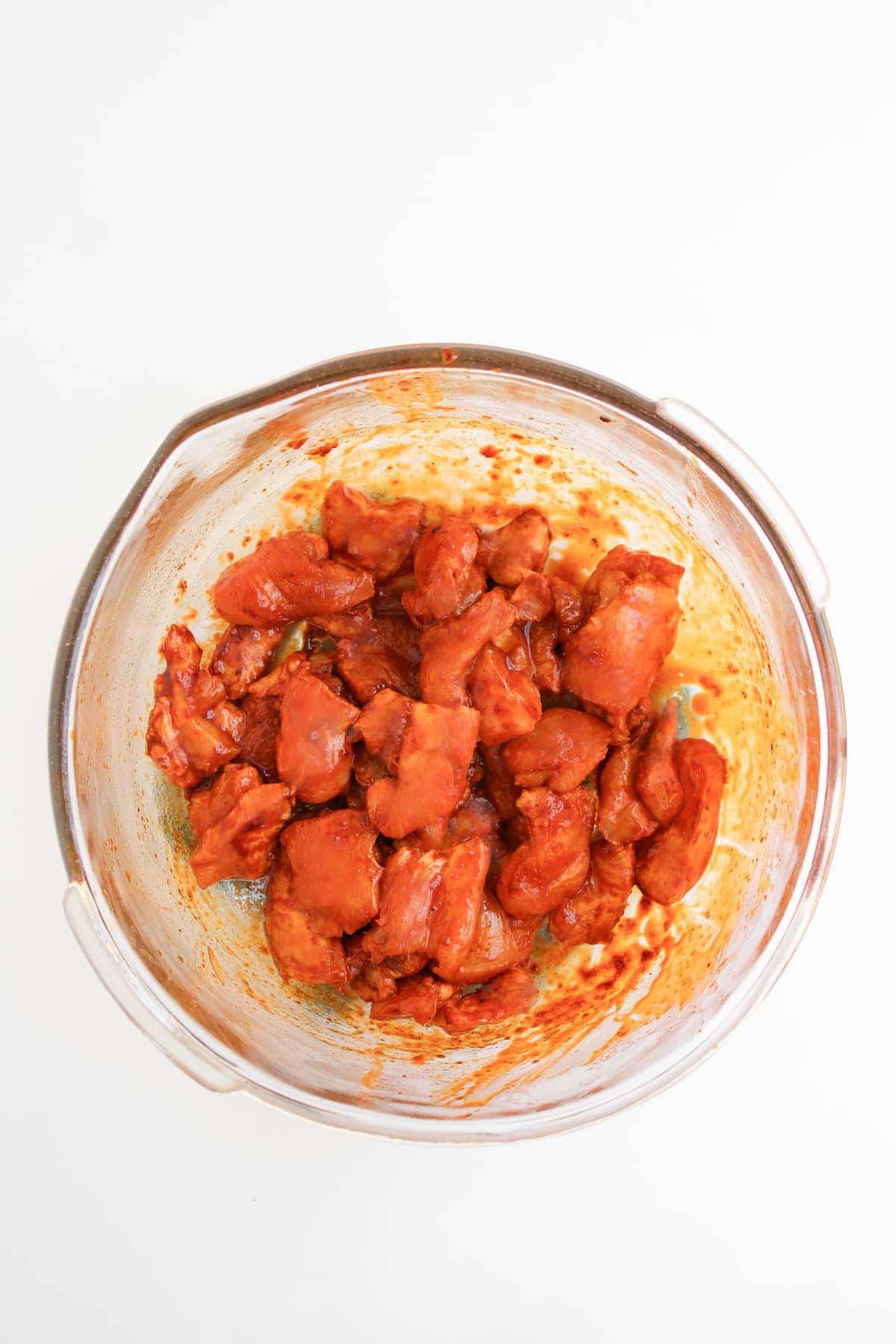 Glass bowl containing raw chicken pieces coated in red marinade, placed on a white background.