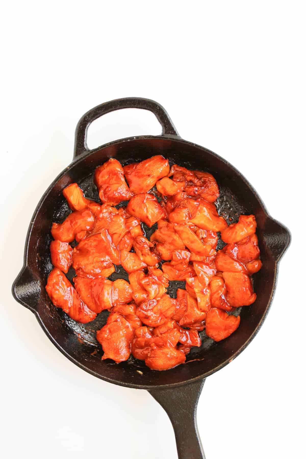 Cooked pieces of reddish-orange marinated chicken in a black cast iron skillet on a white background.