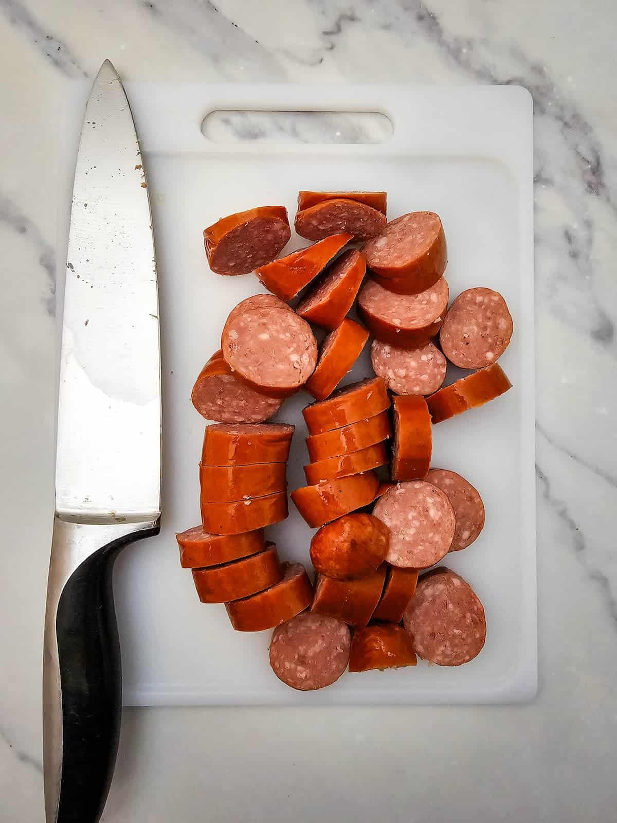 A sliced sausage and a kitchen knife are placed on a white cutting board atop a marble countertop.