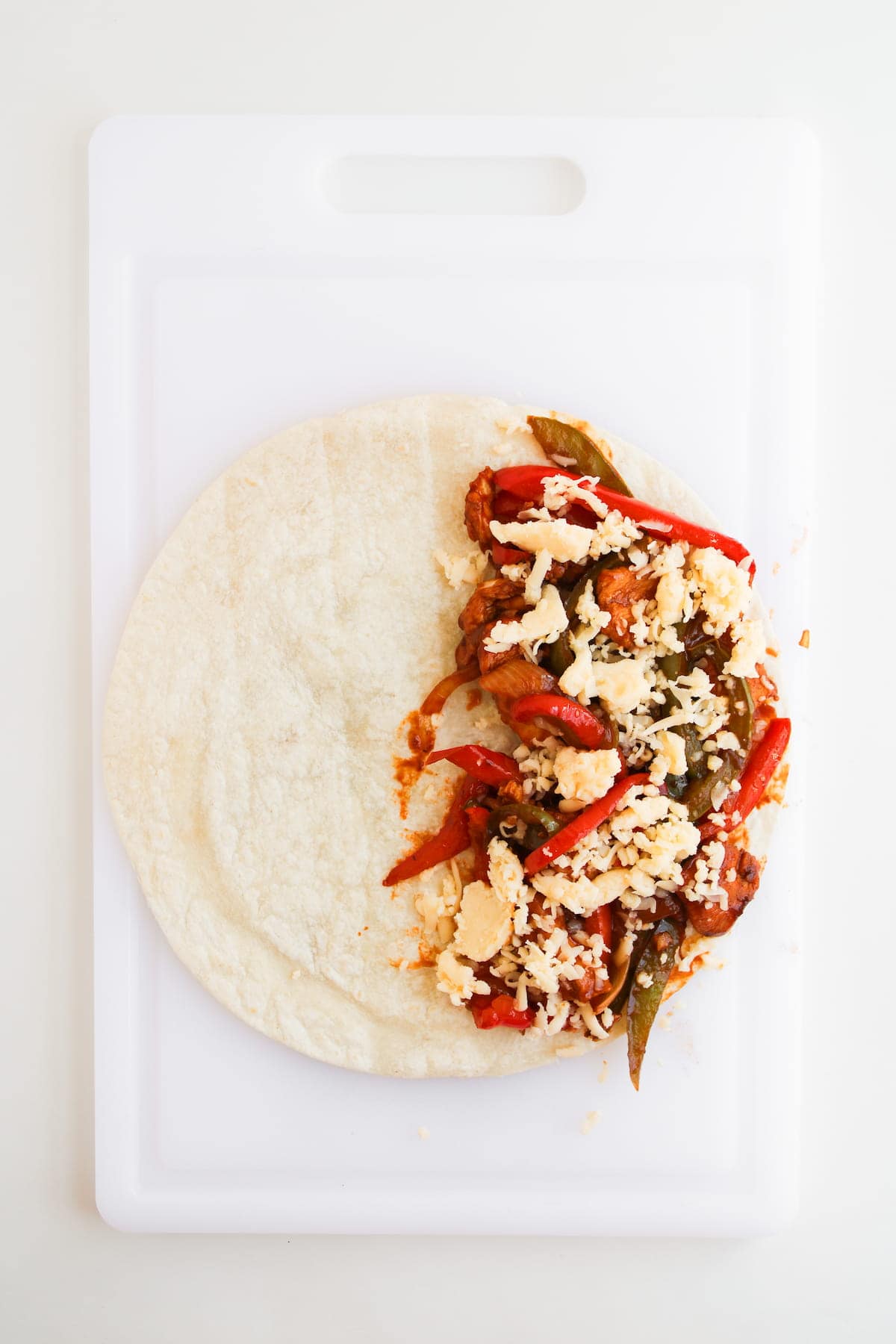 A flour tortilla on a white cutting board, half-filled with sautéed vegetables, tomato sauce, and shredded cheese.