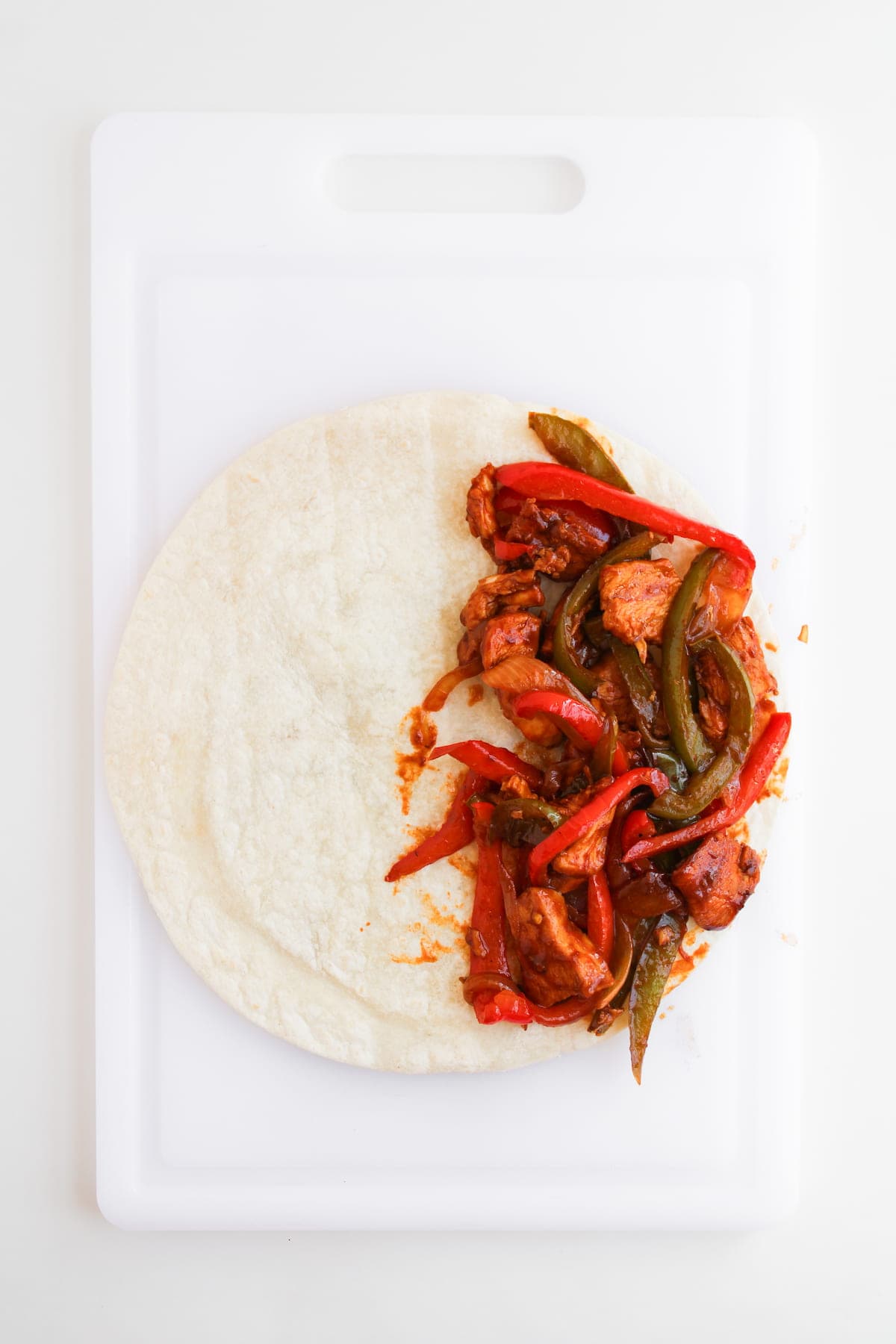 A flour tortilla on a white cutting board, partially filled with cooked chicken, red bell peppers, and green bell peppers.