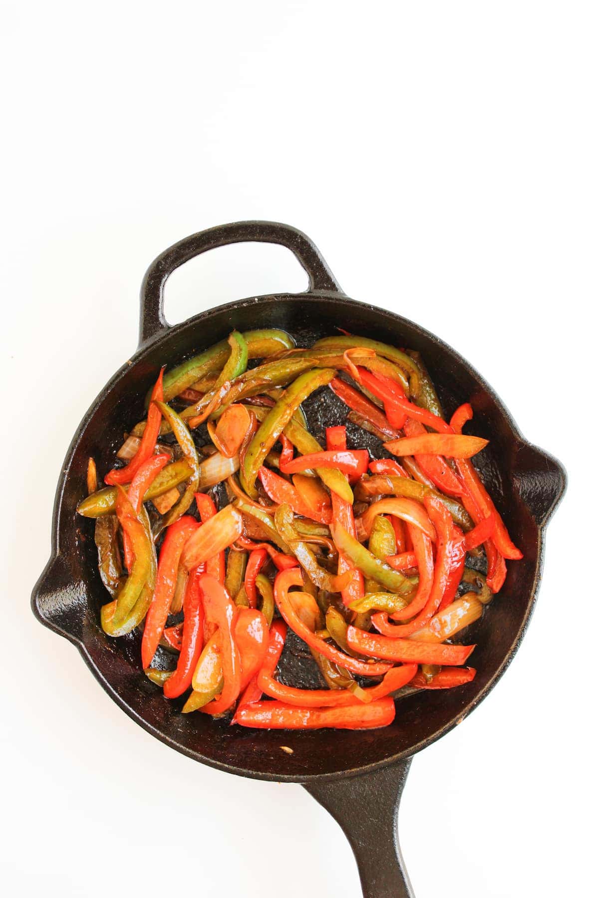 Sliced red and green bell peppers and onions sautéed in a black cast iron skillet on a white background.