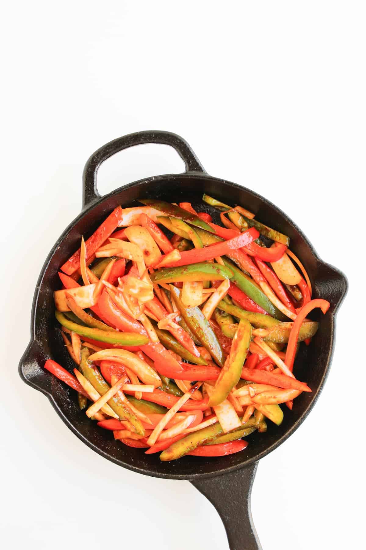 A cast iron skillet filled with cooked sliced bell peppers, onions, and other vegetables on a white background.