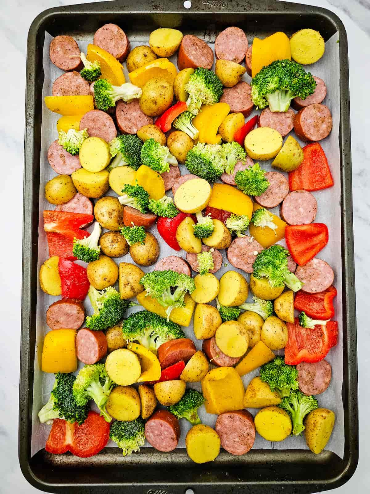Sheet pan filled with sliced sausage, halved baby potatoes, broccoli florets, and chopped red and yellow bell peppers, all arranged on parchment paper.