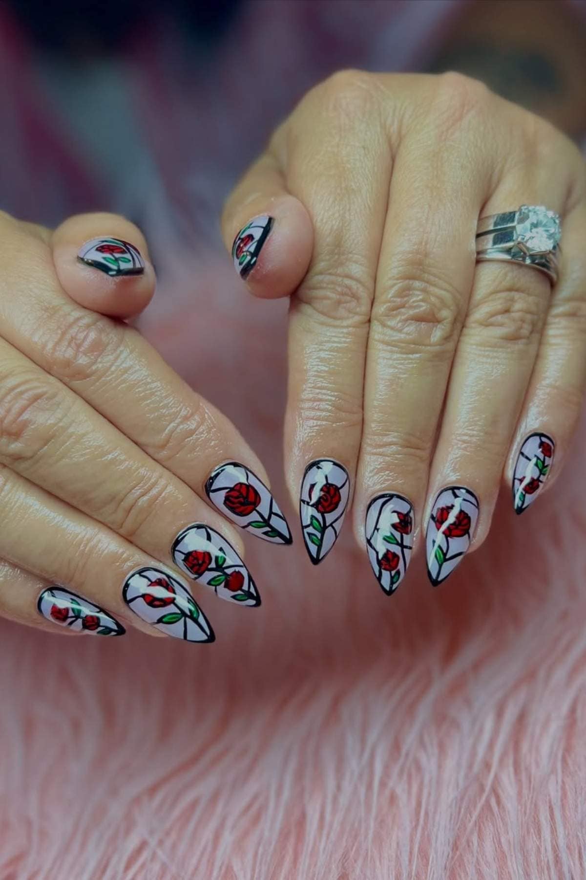 Hands with long, pointed nails featuring detailed red rose nail art outlined in black, resting on a pink textured surface.