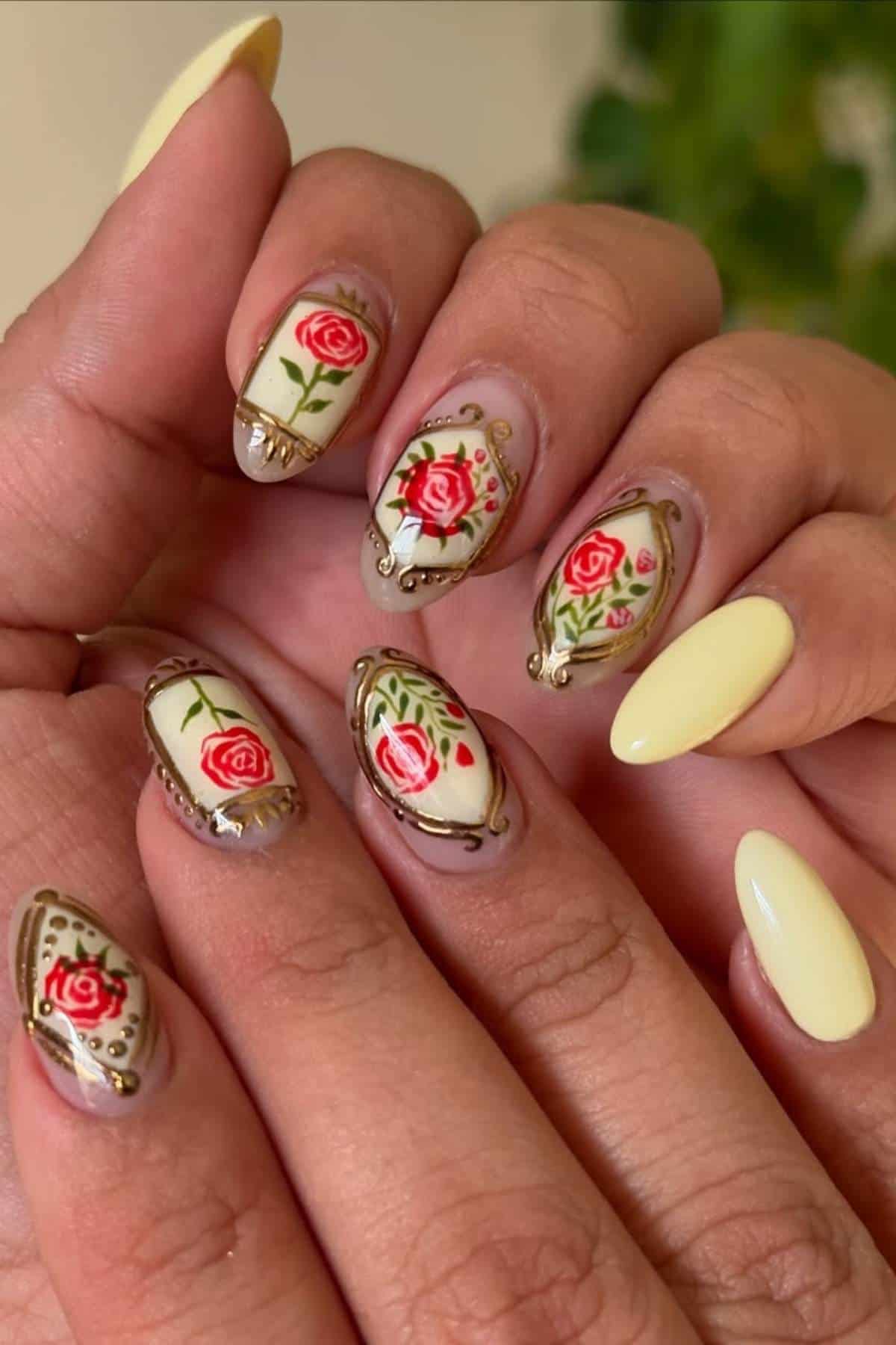 A hand with almond-shaped nails featuring detailed red rose floral designs and gold accents on some nails, and solid pale yellow polish on others.