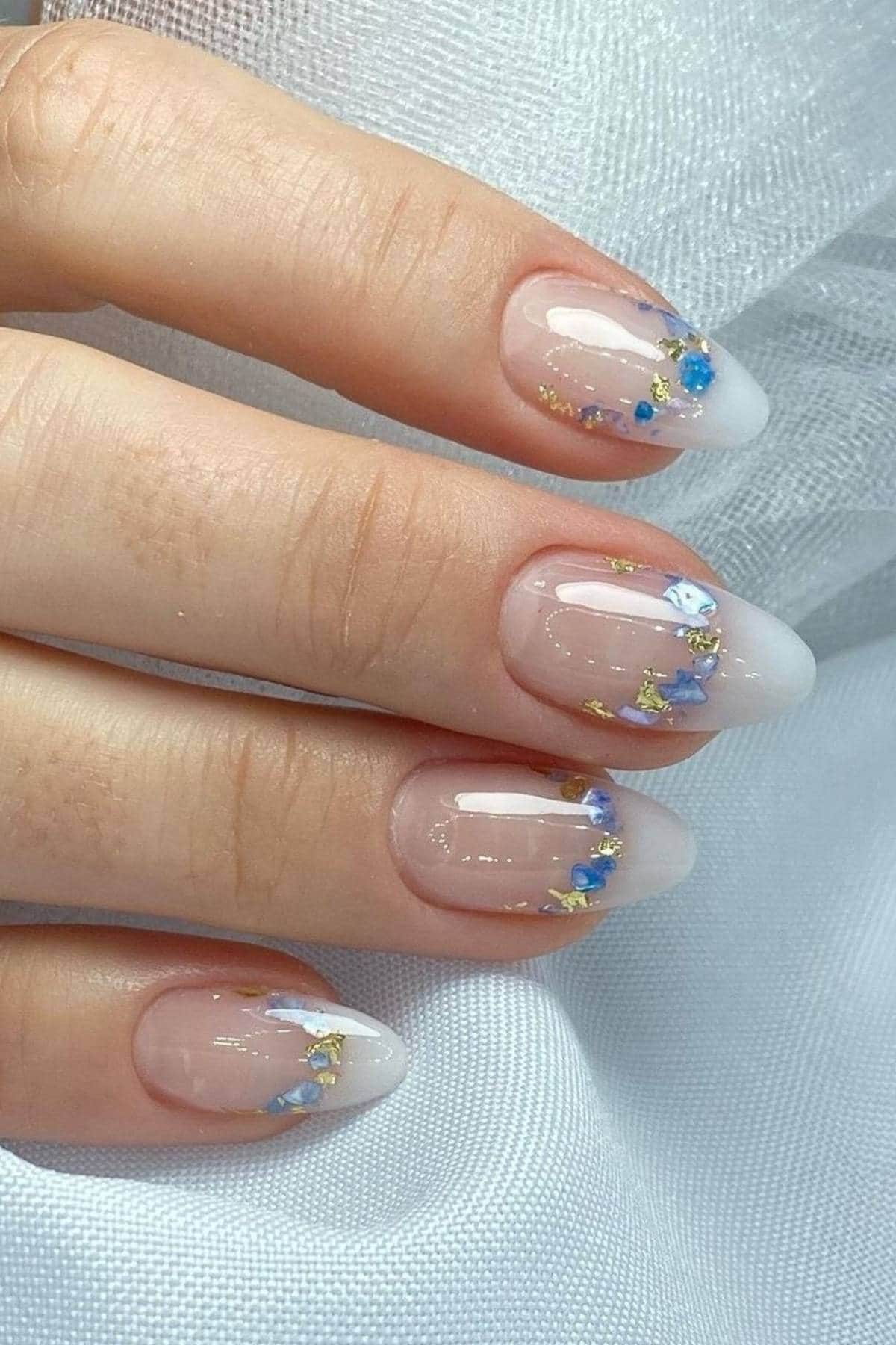 A hand with almond-shaped nails featuring a translucent pink base, white ombre tips, and blue, gold, and white floral accents on each nail.