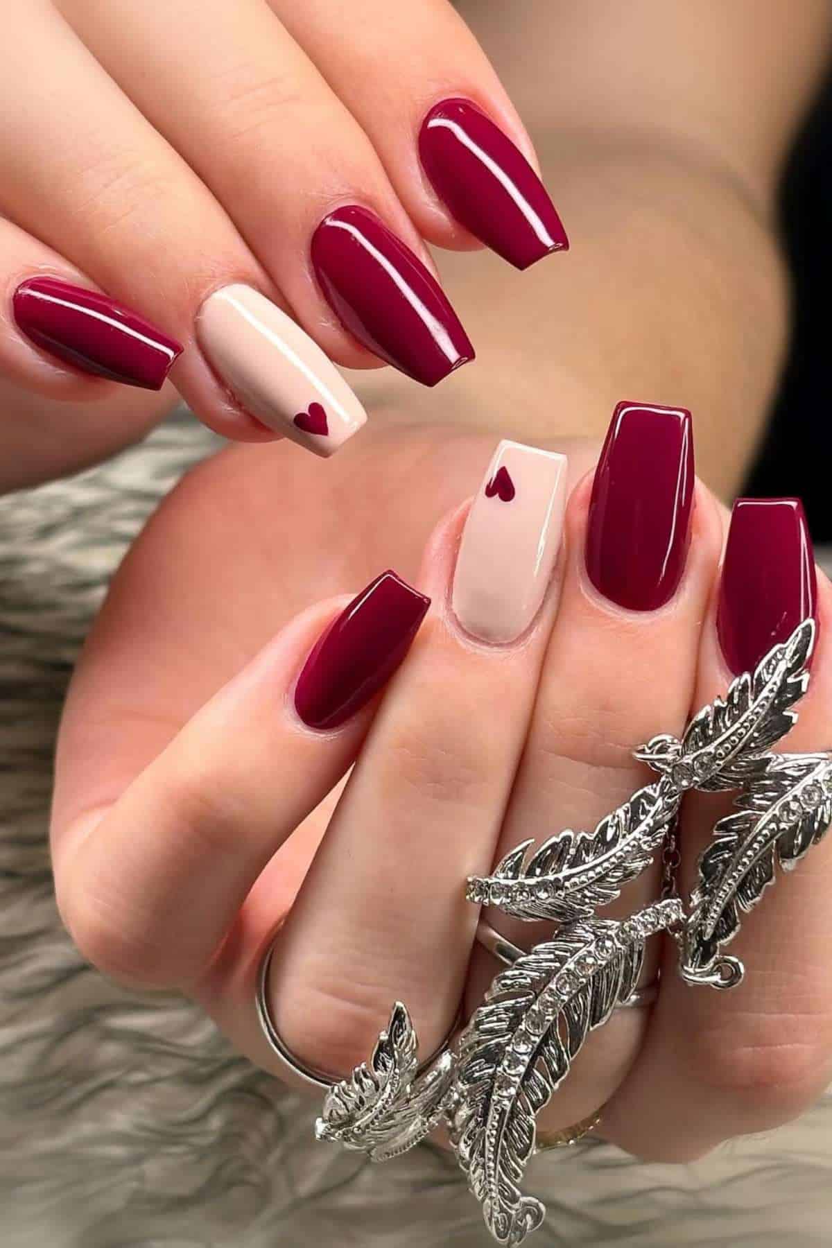 Hands with long, square-shaped nails painted burgundy and light pink, featuring small heart designs on the ring fingers; wearing a silver feather ring.