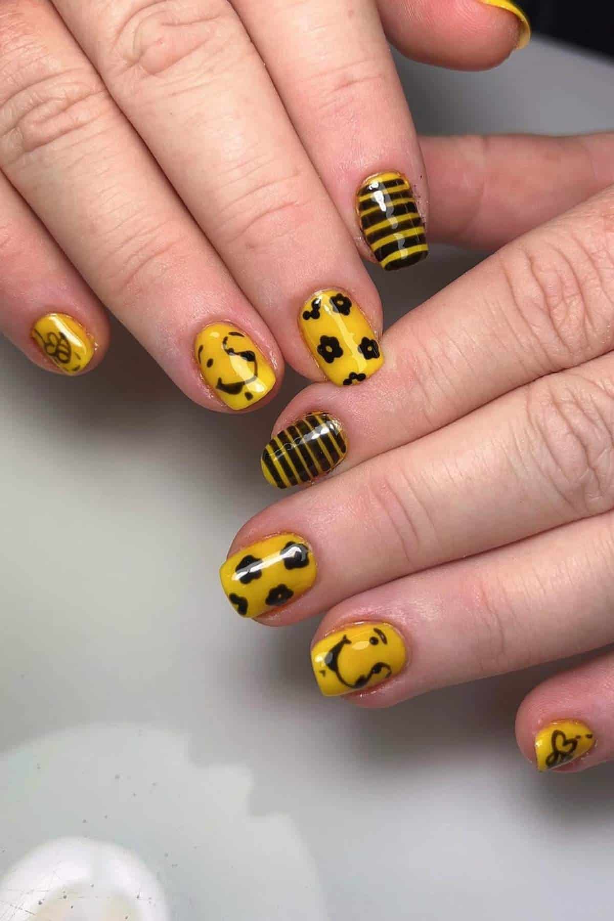 Close-up of hands with yellow nail polish featuring black floral designs, stripes, polka dots, a smiley face, and a small bee on short, neatly manicured nails.
