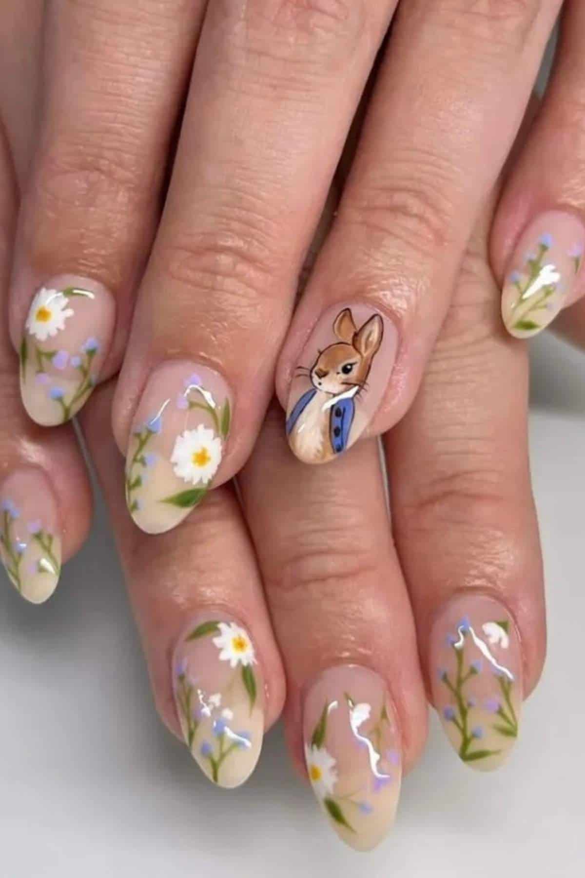 Hands with almond-shaped nails painted with white daisies and green stems; one nail features a detailed illustration of a rabbit wearing a blue jacket.