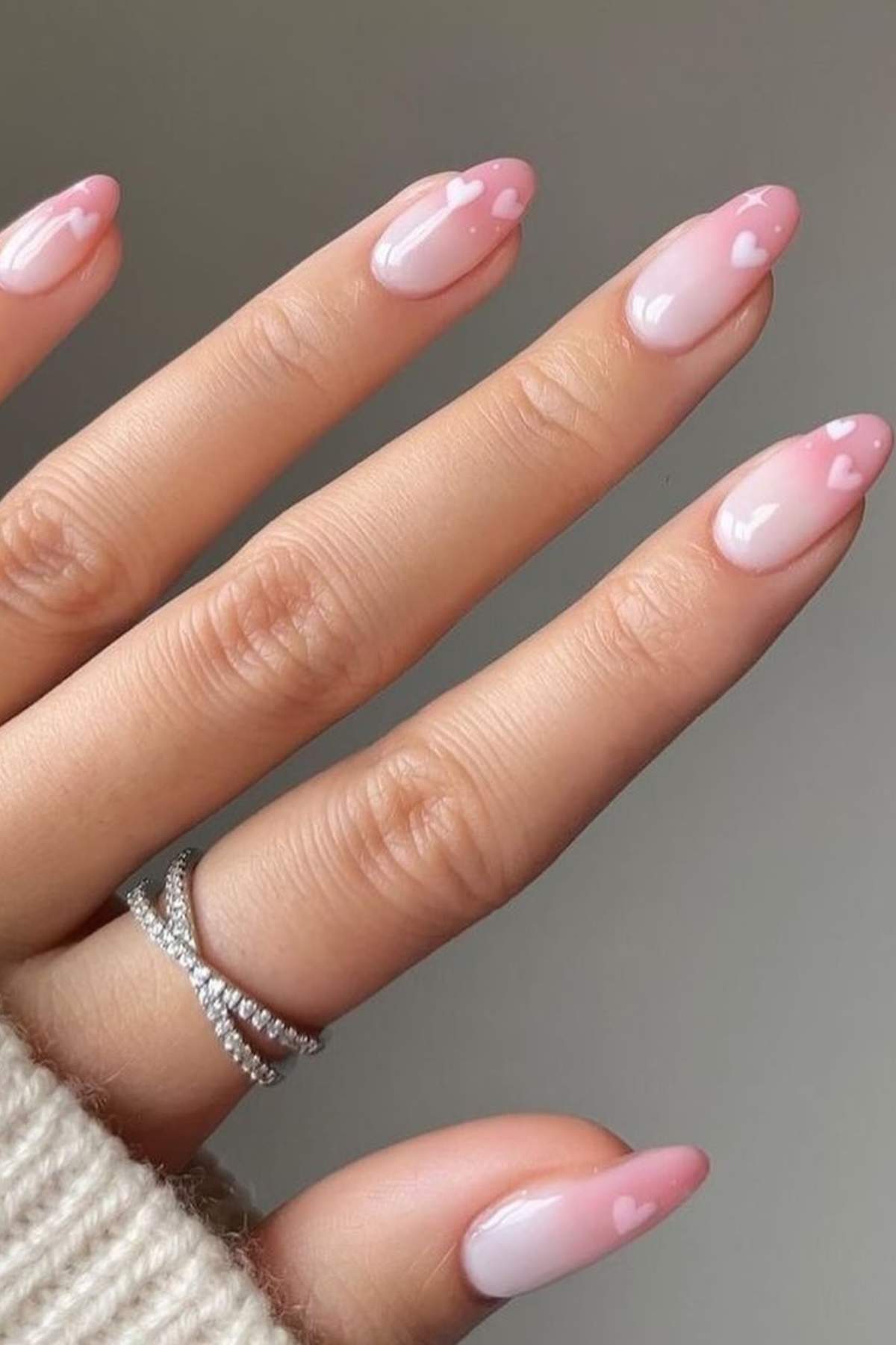 A hand with almond-shaped nails painted in pale pink with small white heart designs near the tips. The person is wearing a silver ring and a cream-colored sweater.