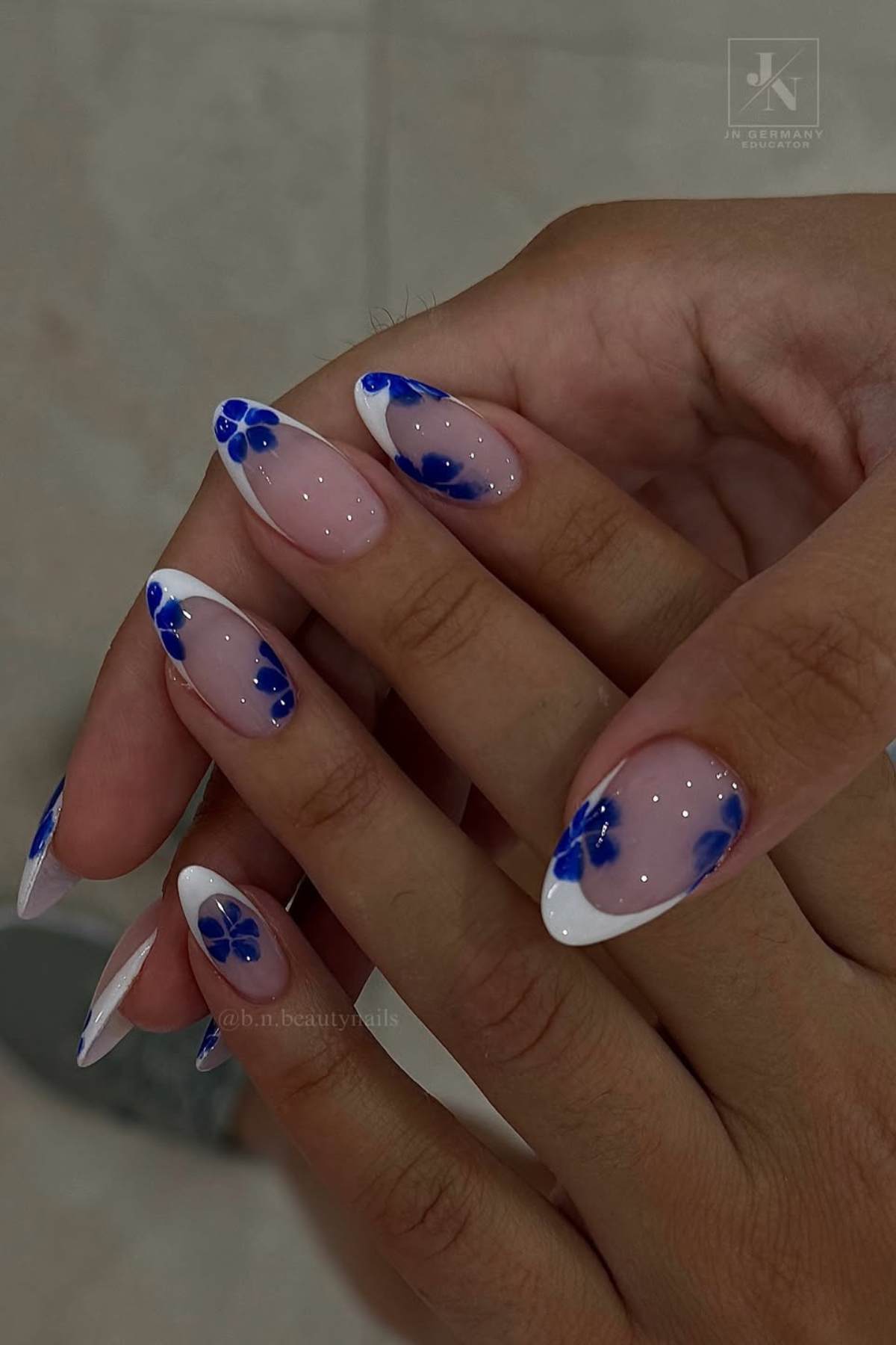 Close-up of hands with almond-shaped nails featuring a French tip design and blue flower nail art on a light pink base.