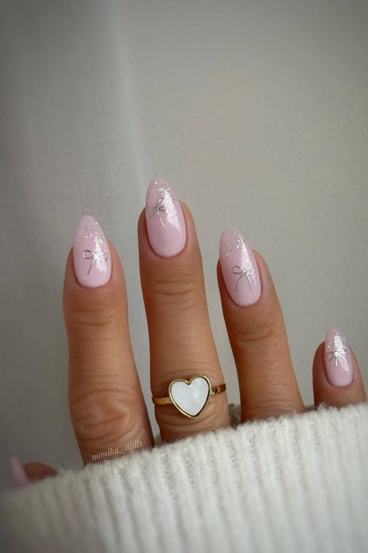 A hand with almond-shaped, pale pink nails featuring white bows and glitter accents, wearing a gold ring with a heart-shaped white stone.