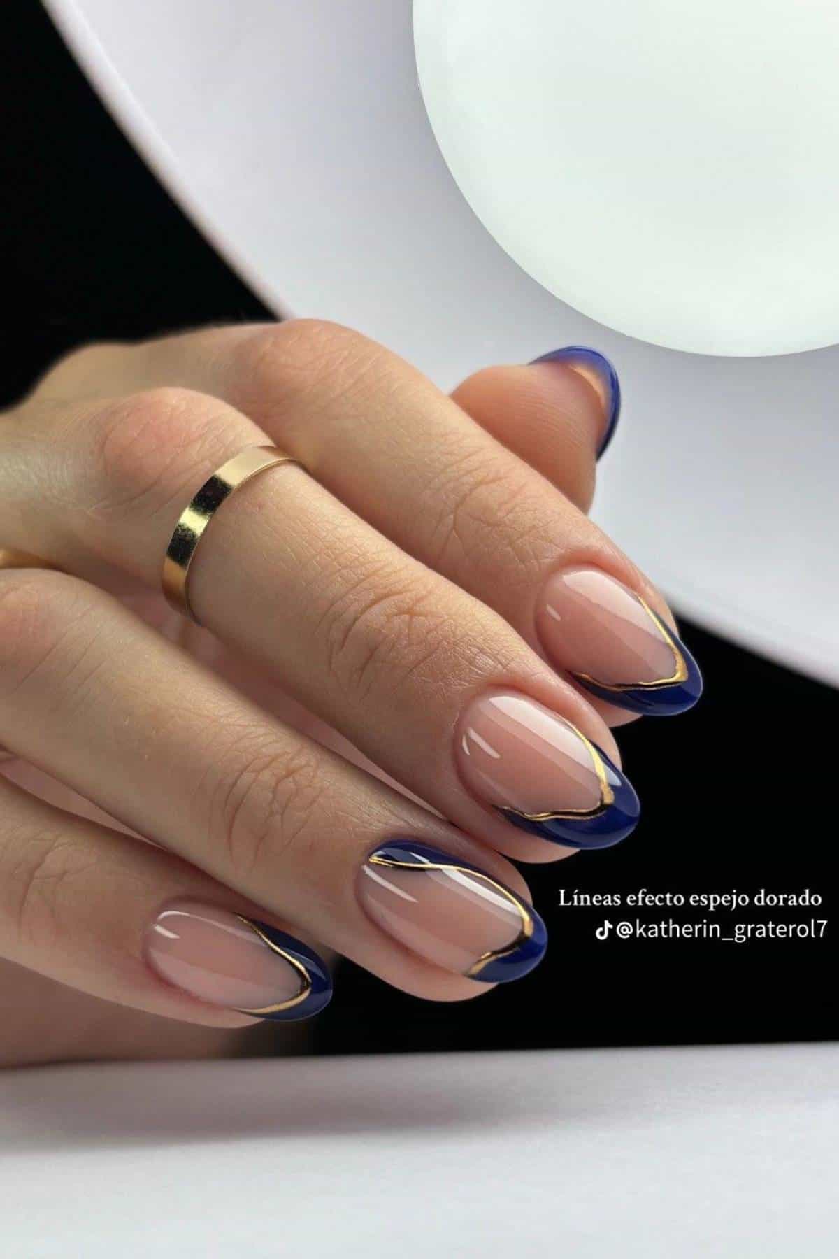 A hand with almond-shaped nails featuring a glossy nude base, gold mirror effect lines, and dark blue tips, wearing a simple gold ring.