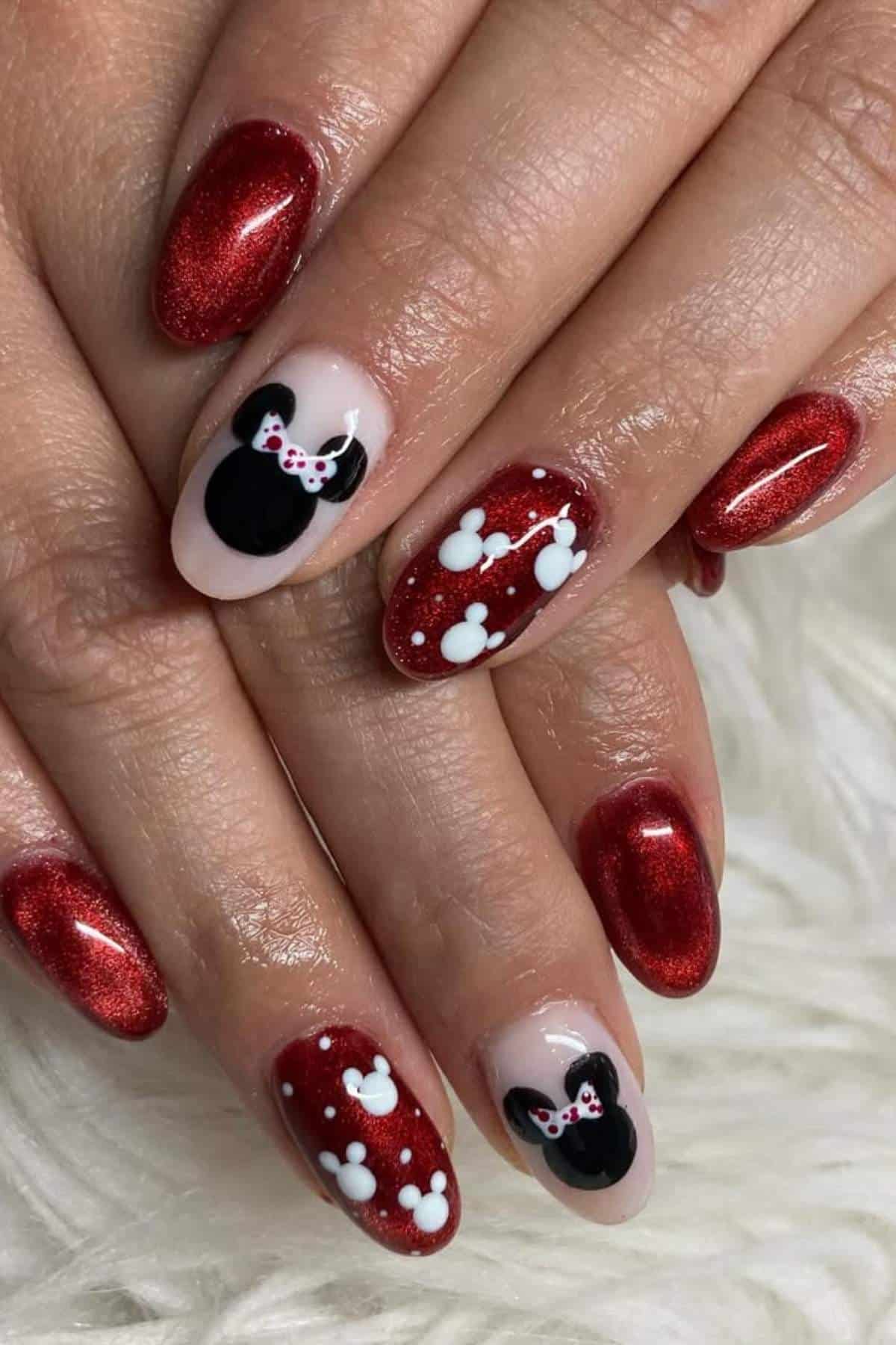 Hands with red glittery nails featuring Minnie Mouse head designs and white polka dots on accent nails, on a white textured surface.