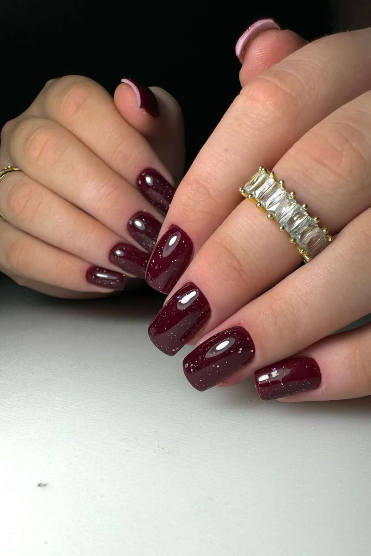Hands with dark red, glossy square-shaped nails featuring subtle glitter; one hand wears a gold ring with rectangular clear stones.
