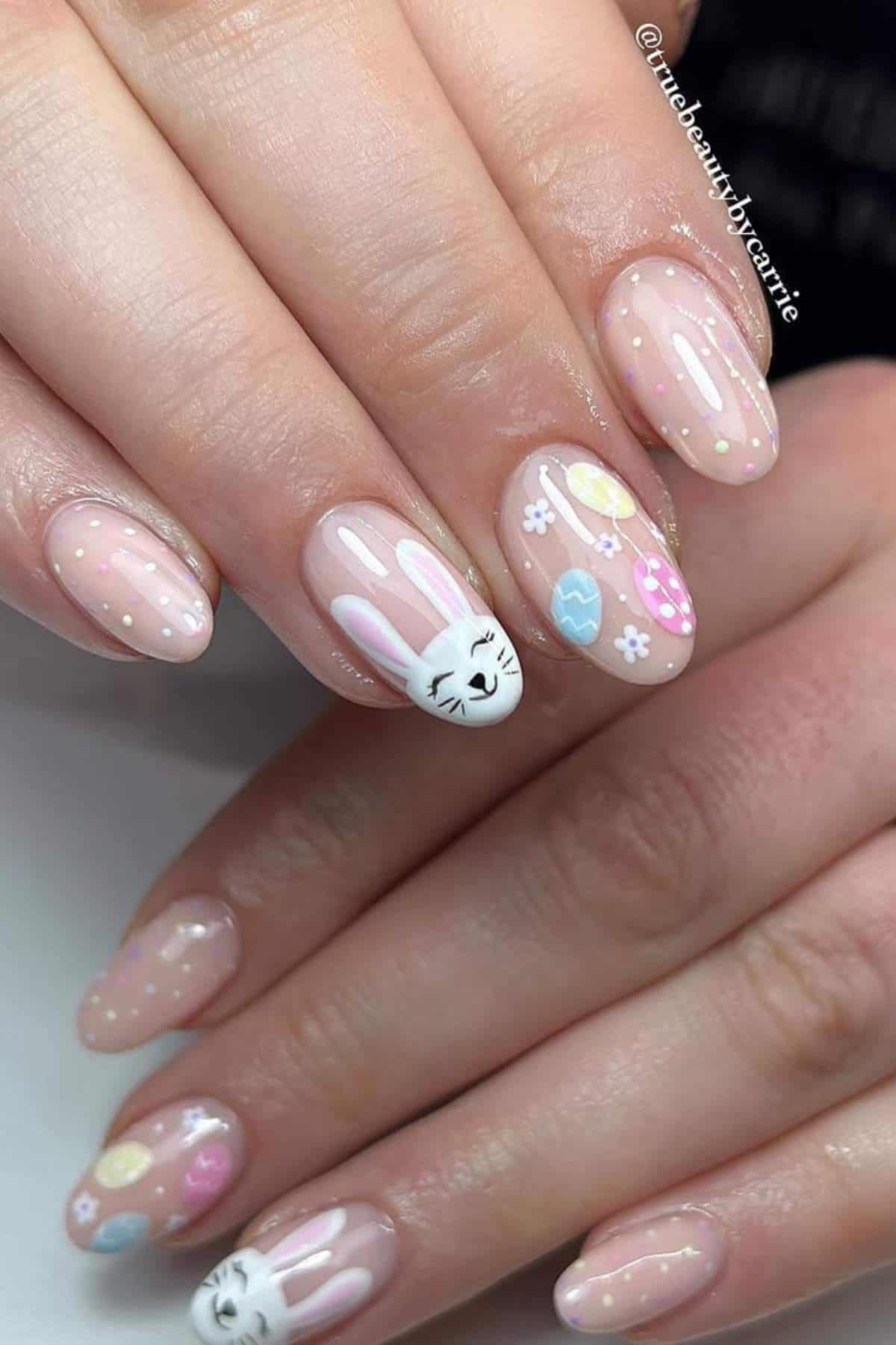Close-up of hands with nude nails featuring white polka dots, pastel Easter egg designs, and a white bunny face on accent nails.