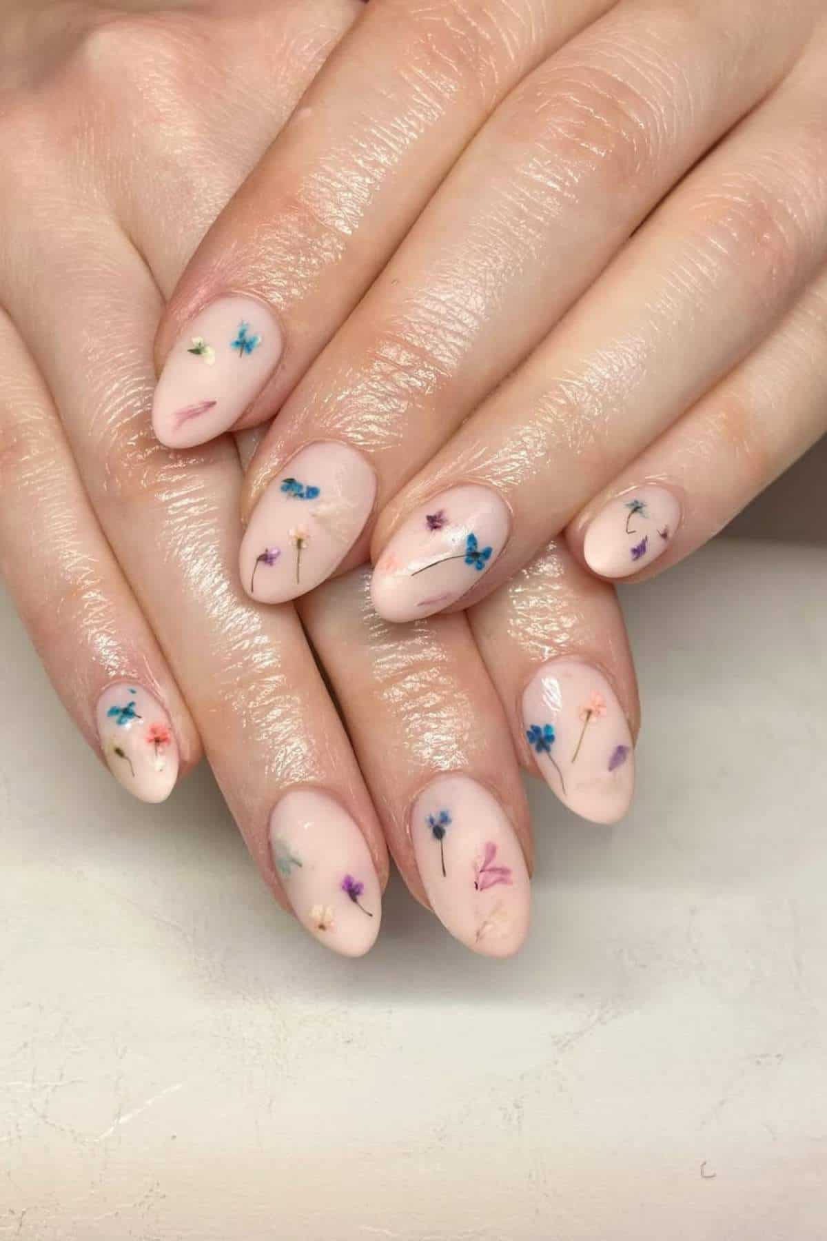 Hands with almond-shaped nails painted in a pale nude shade, each nail featuring small, colorful dried flower designs.