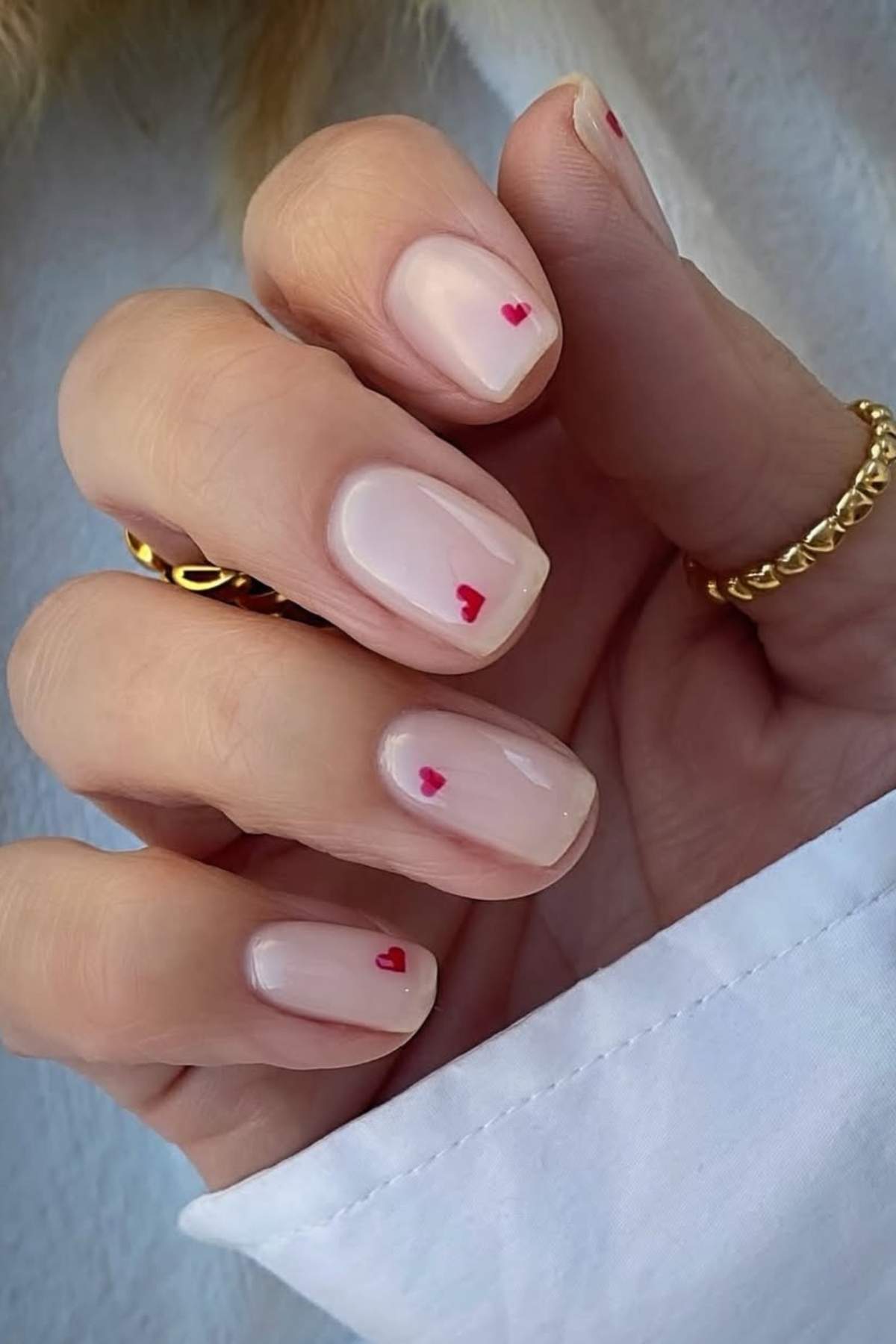 A hand with short, pale pink manicured nails, each featuring a small red heart near the cuticle. The hand is wearing gold rings and a white sleeve is visible.