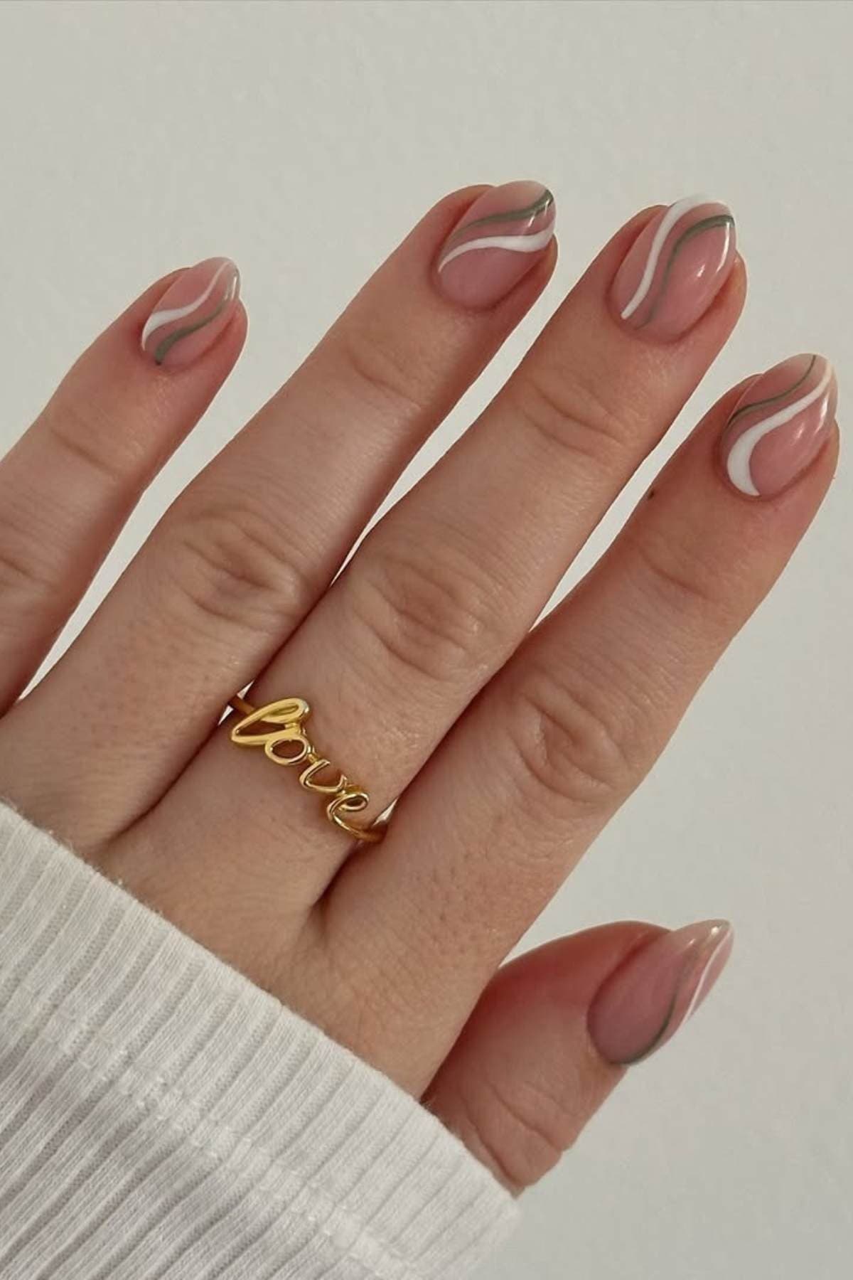 A hand with nude-colored nails featuring pink, white, and gray abstract wavy designs; the person wears a gold "love" ring and a white long-sleeve top.