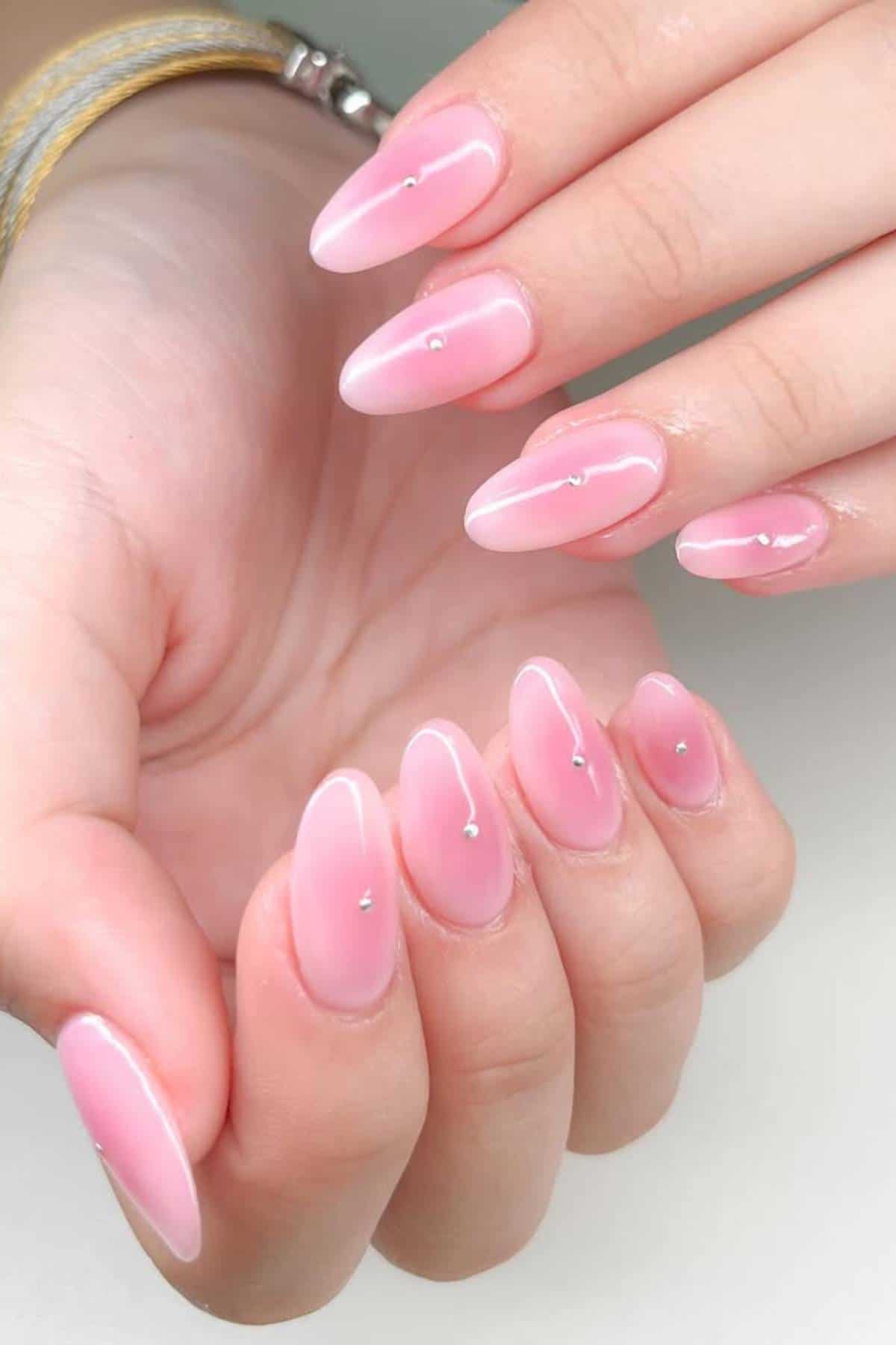 A hand with long, almond-shaped nails painted pink with a glossy finish and small silver studs at the center of each nail.