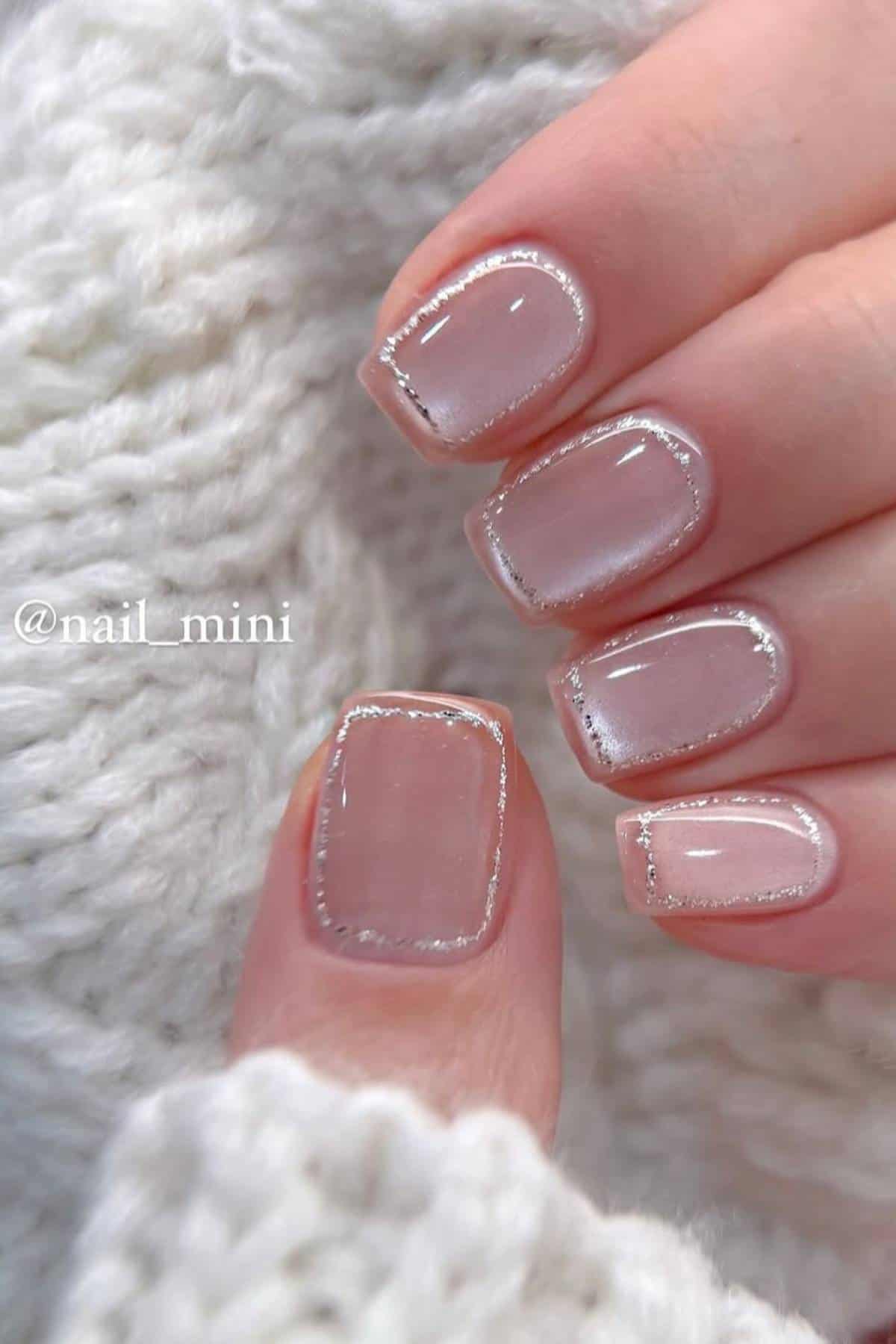A hand with short, square-shaped nails featuring a sheer nude polish and fine silver glitter outlining the edges, resting on a white knitted fabric.