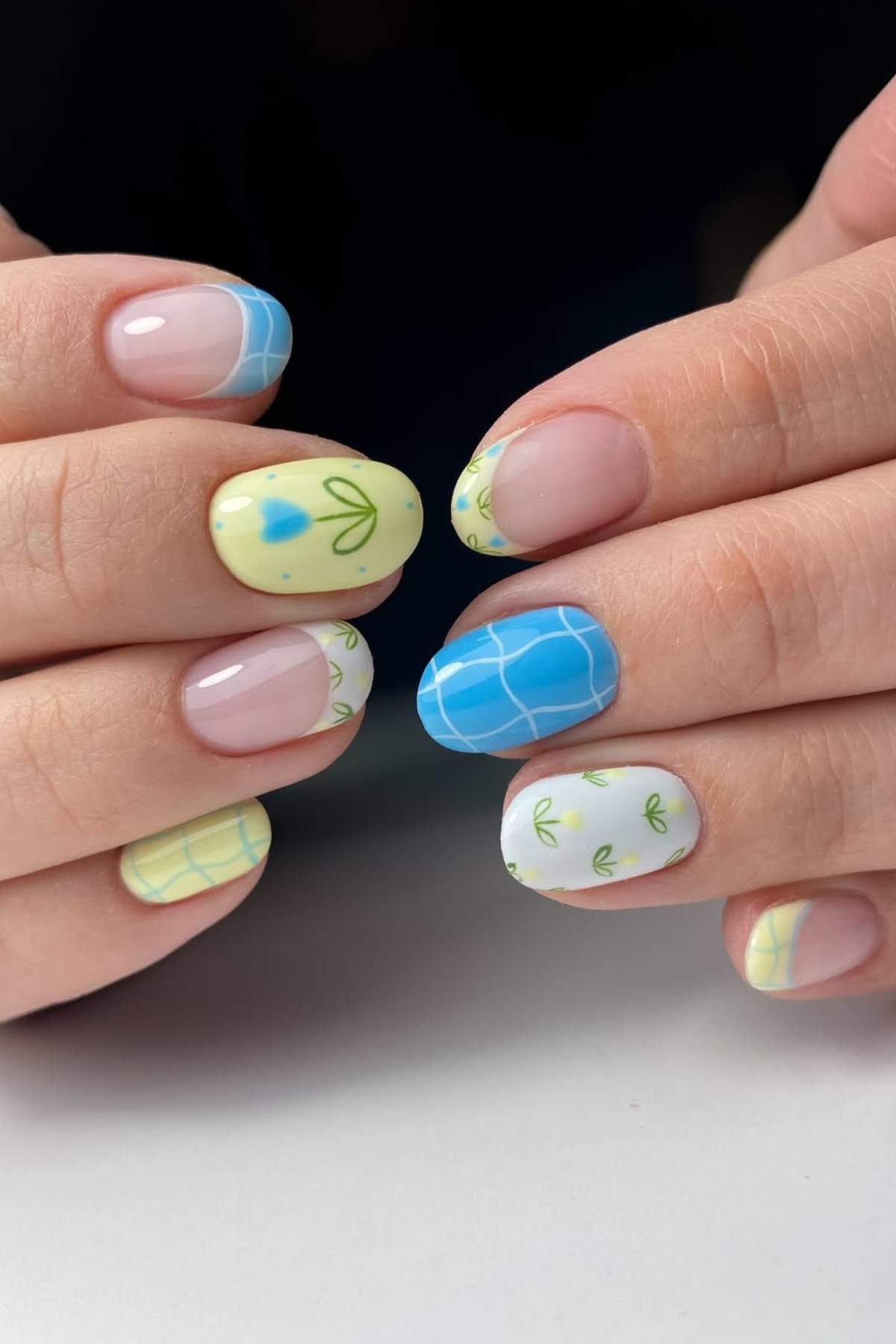 Hands with manicured nails featuring blue, yellow, and white nail polish, decorated with grid patterns, hearts, and small green leaf designs.
