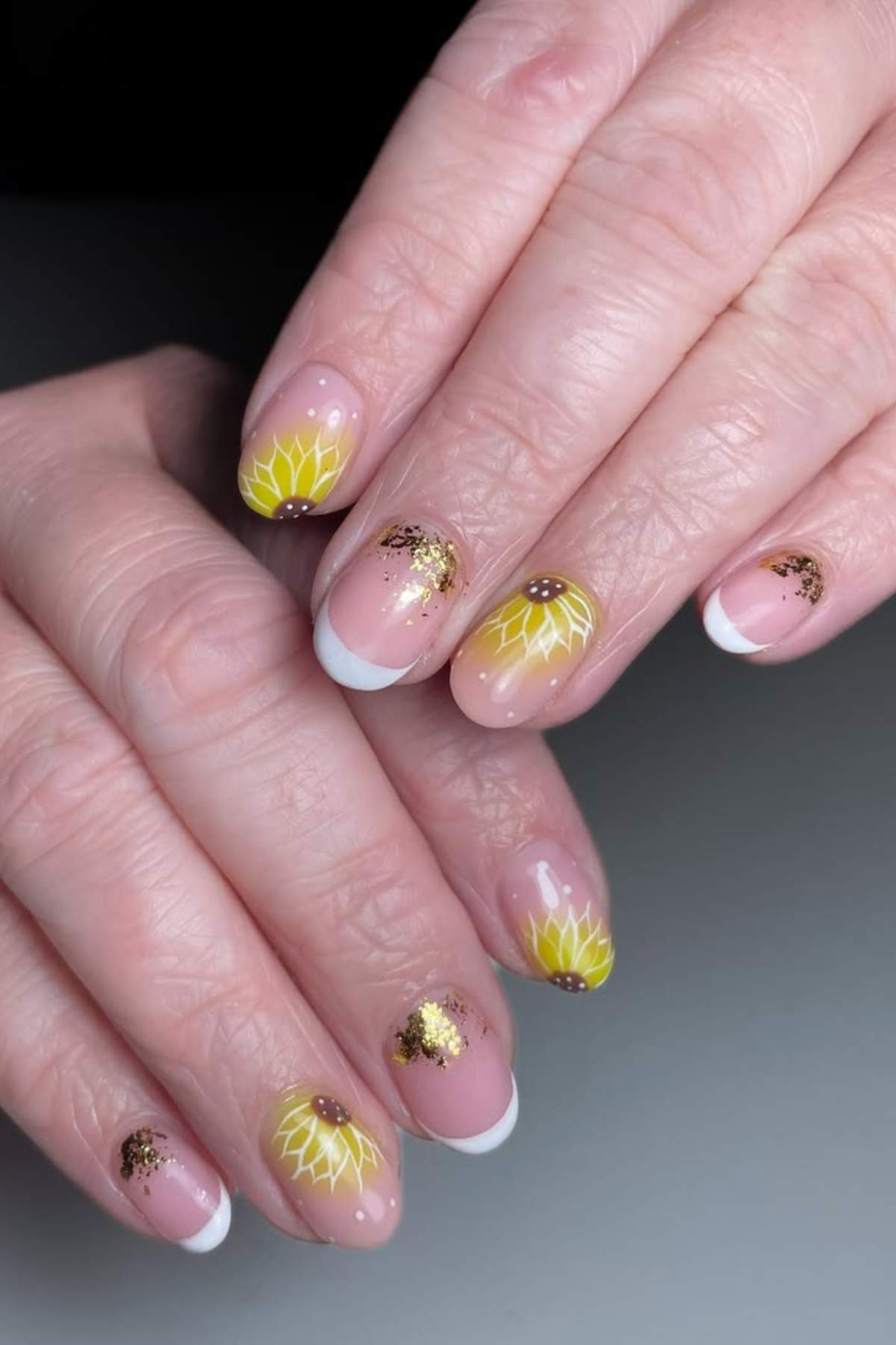 Hands with manicured nails featuring yellow sunflower designs, gold glitter accents, and white French tips on a natural pink base.