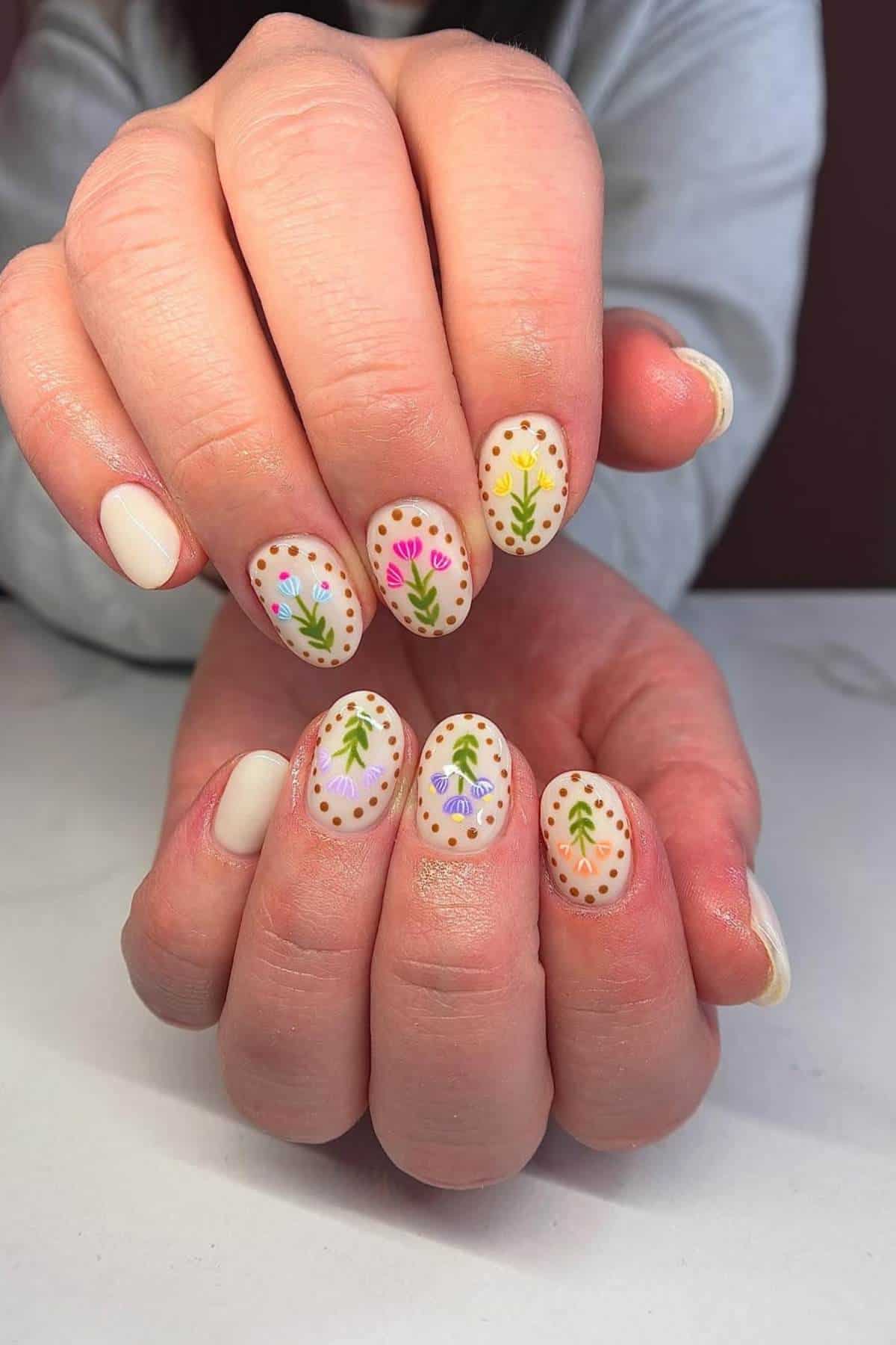 Close-up of hands with oval-shaped nails, each painted with a white base and decorated with colorful floral and leaf designs bordered by small brown dots.