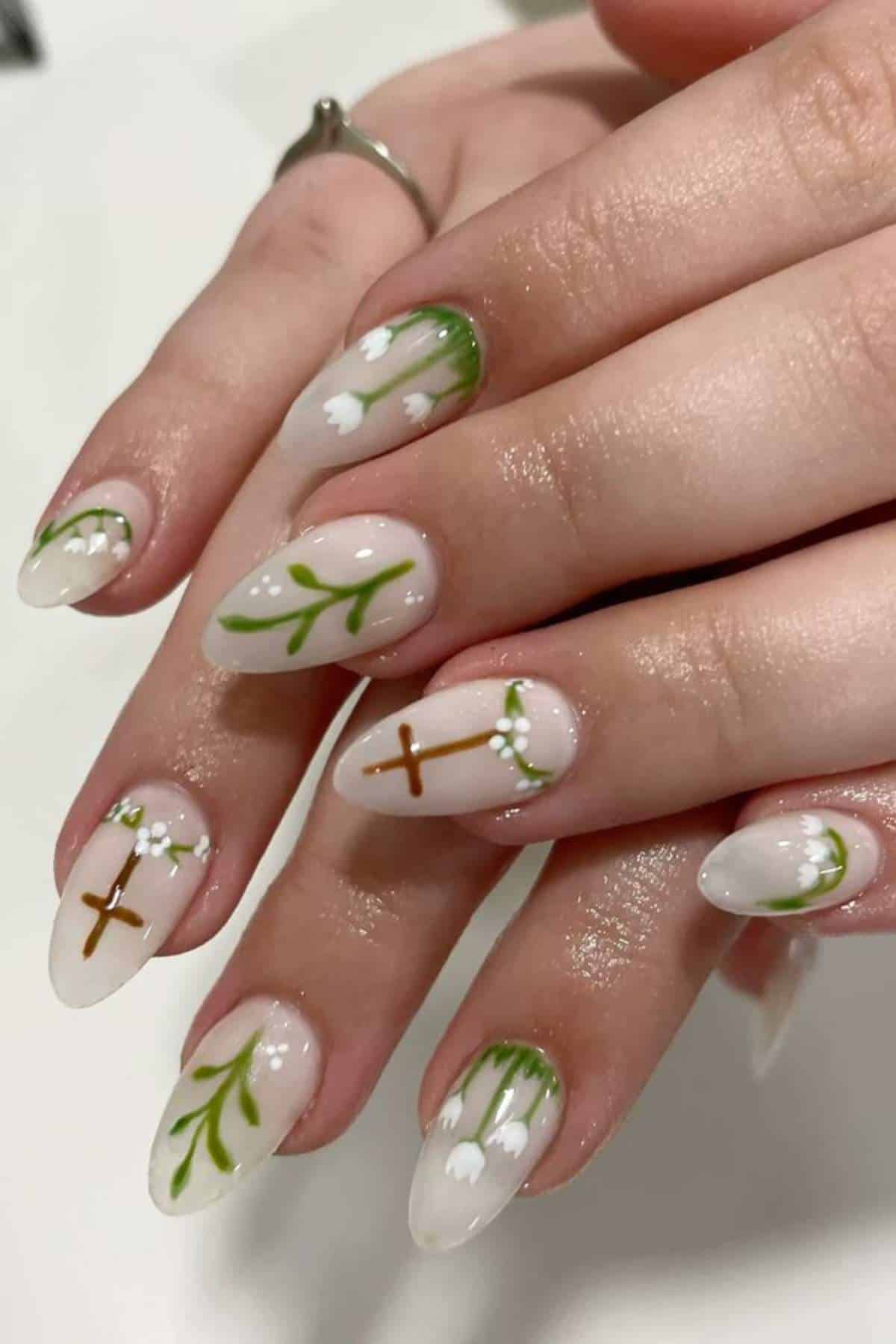 Close-up of hands with almond-shaped nails featuring green leaves, white flowers, and brown crosses painted on a nude base.