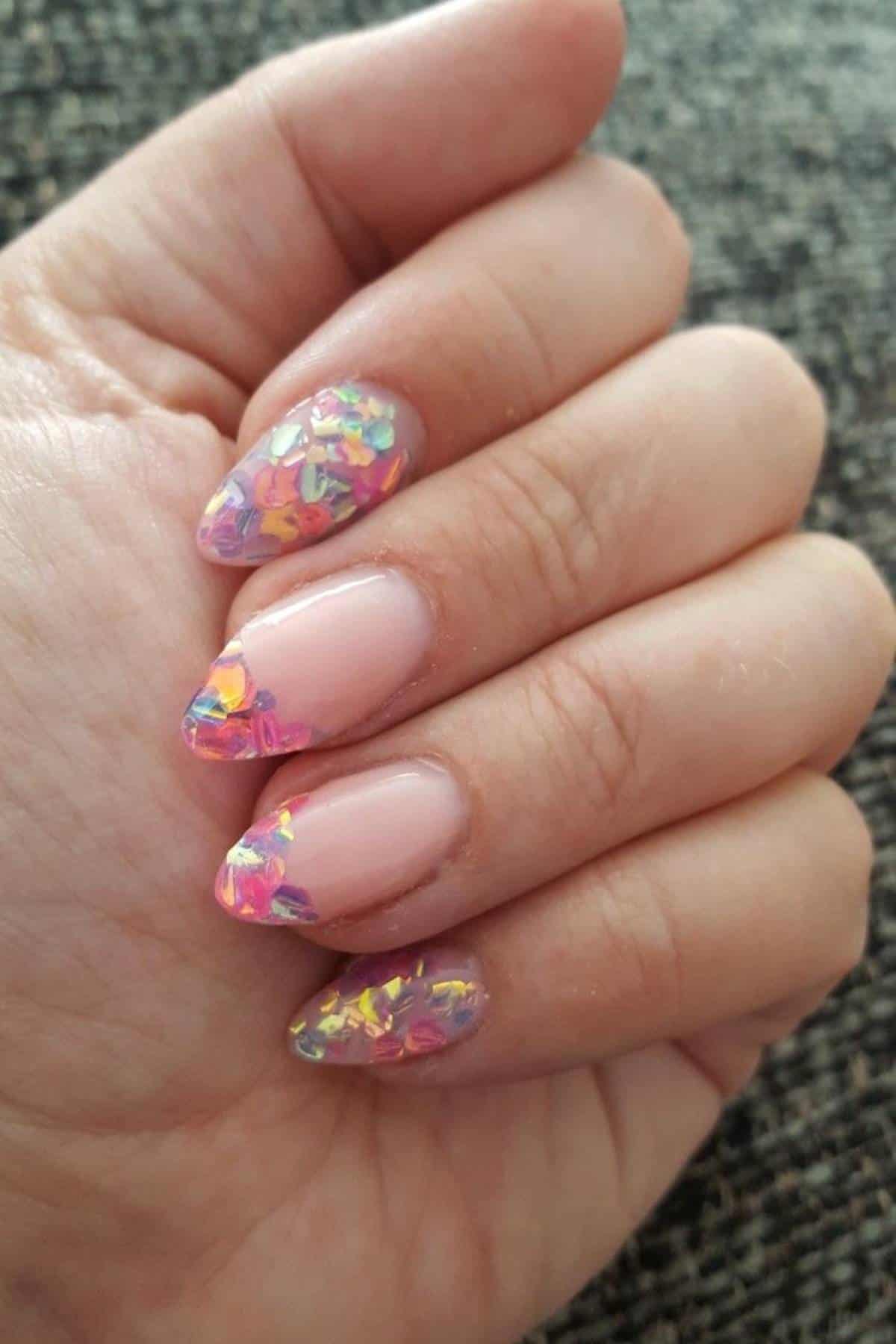 A hand with almond-shaped nails featuring a nude base and colorful, iridescent foil tips.