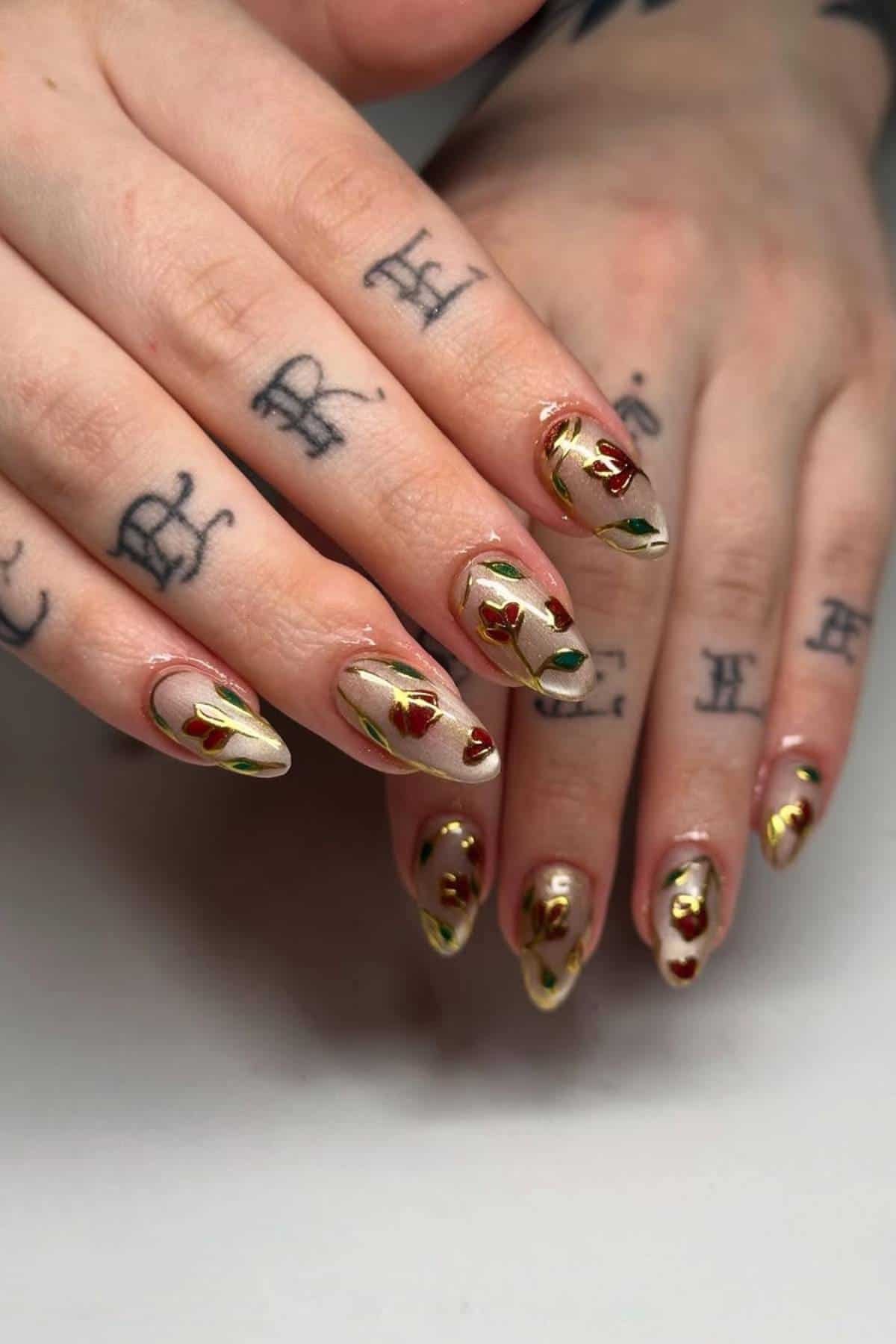 Hands with tattoos on the fingers show almond-shaped nails painted with a beige base and decorated with red rose designs and gold accents.