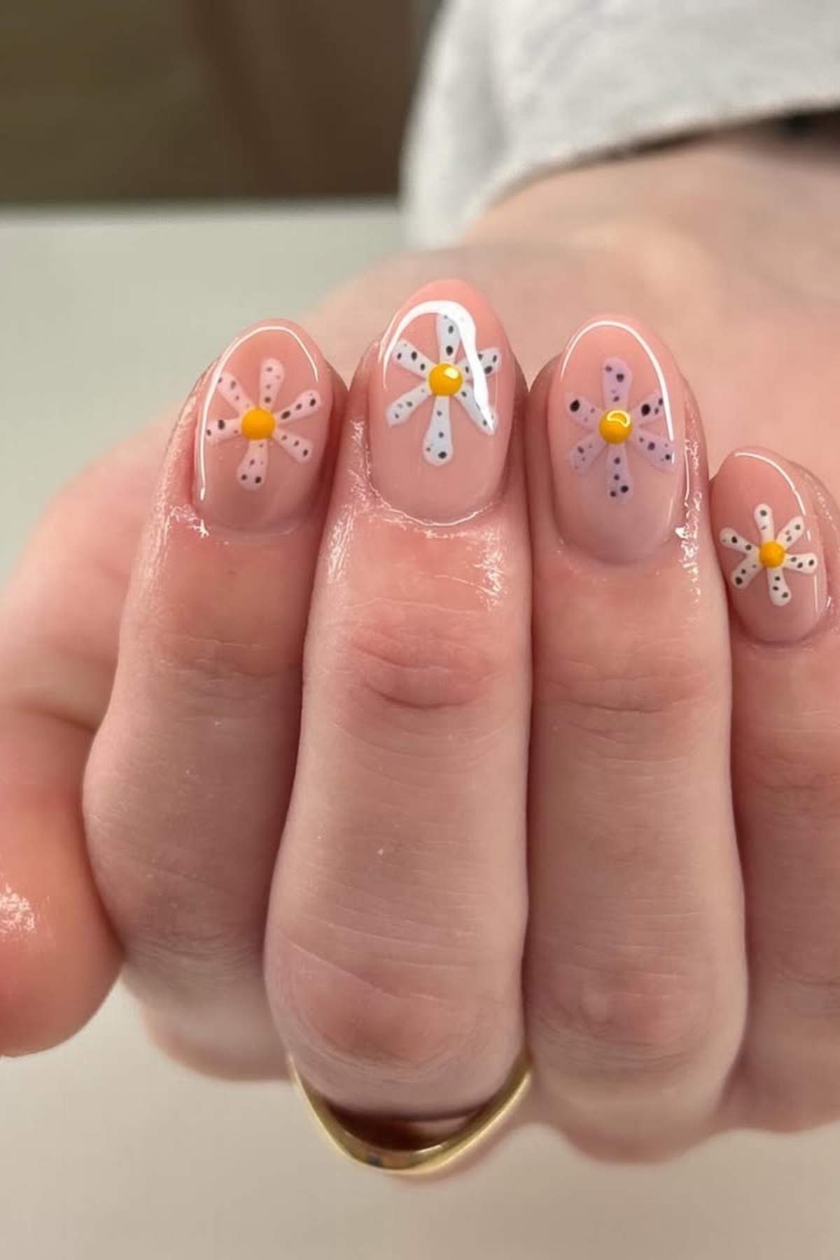 A hand with short natural nails painted with a nude polish and decorated with pastel flower designs, each with a yellow center and dotted petals.