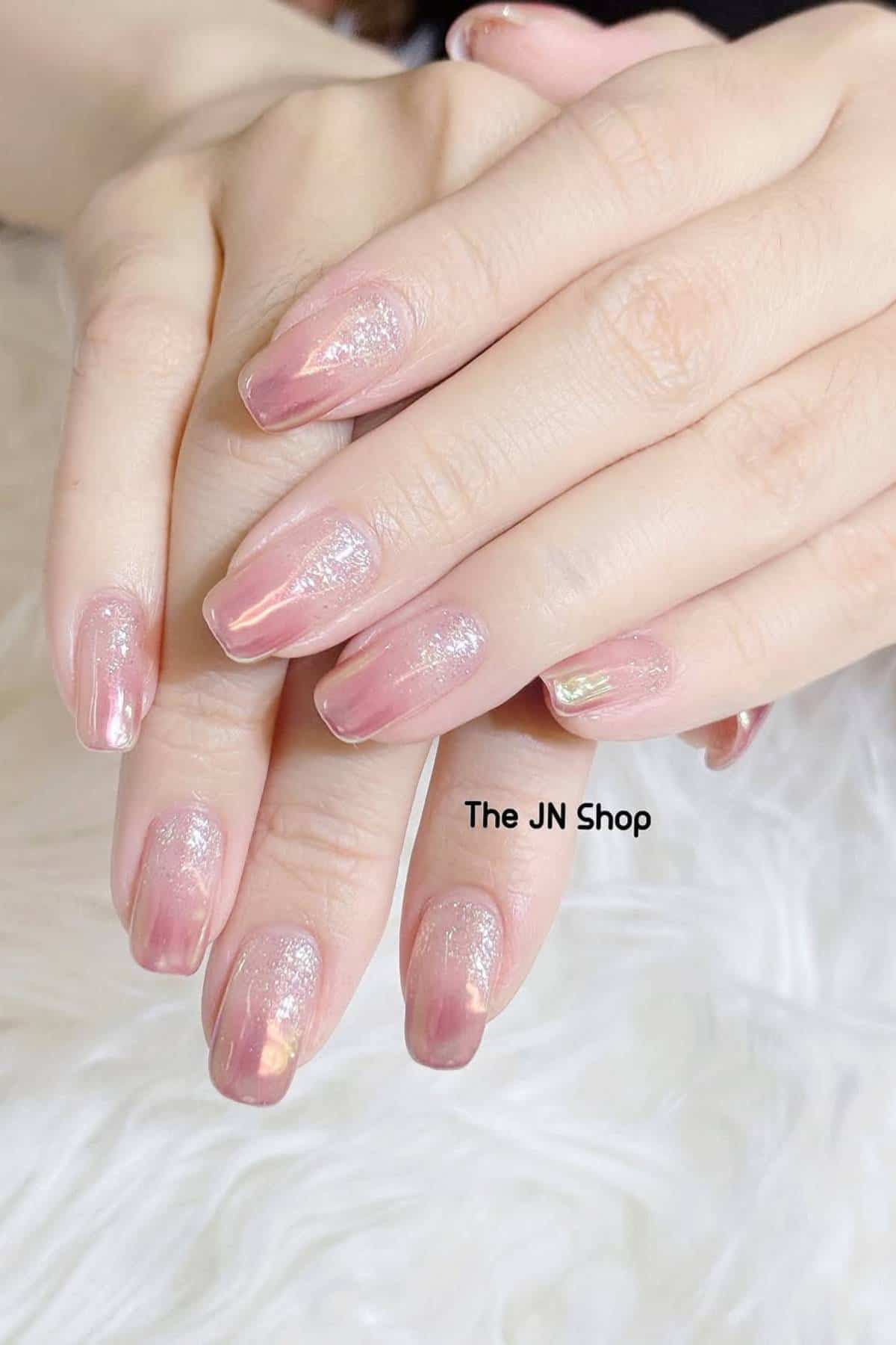 Close-up of hands with medium-length, glossy pink nails featuring a subtle shimmer and iridescent effect, displayed on a soft white surface.