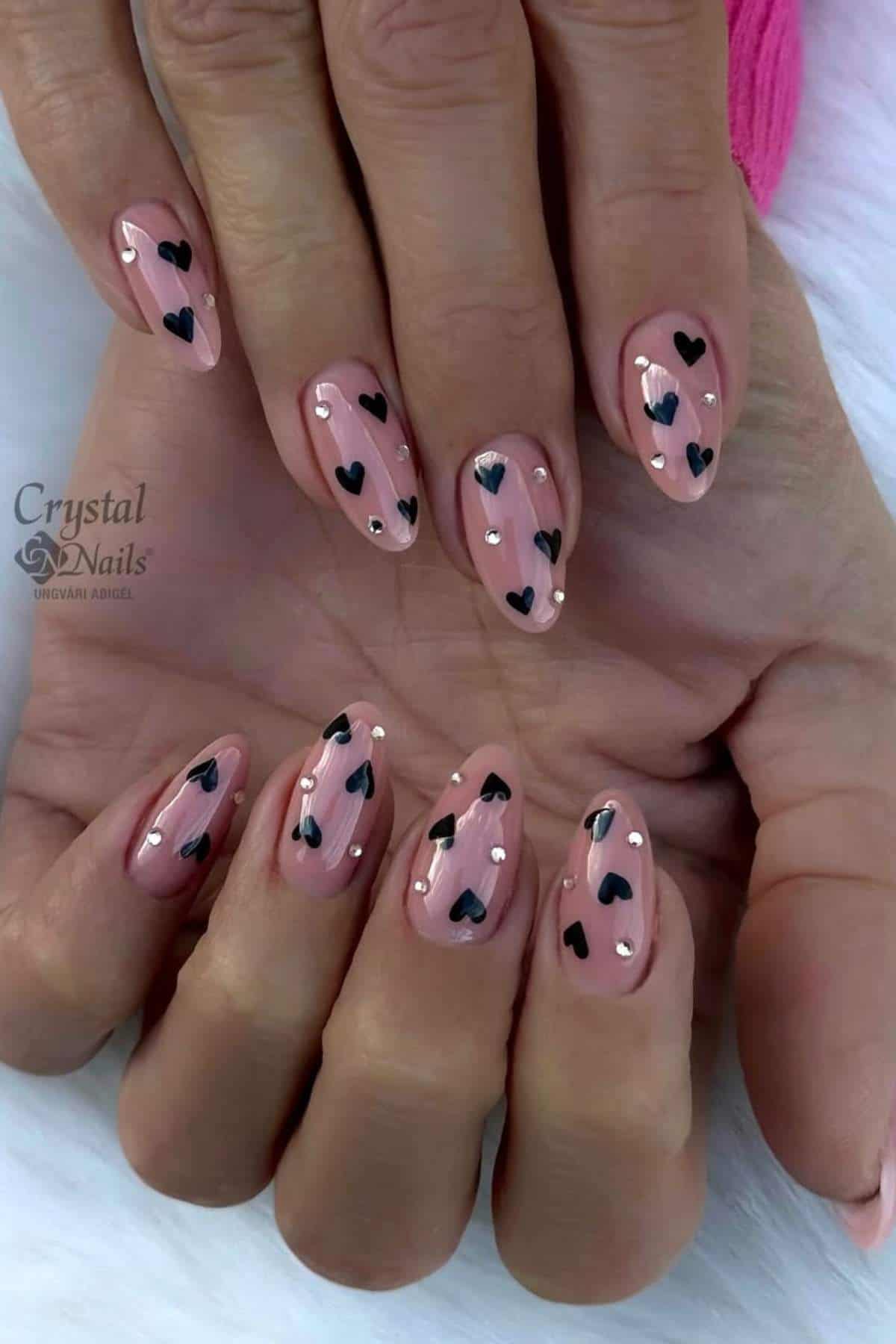 Hands with almond-shaped nails painted nude pink, decorated with small black hearts and silver dots on each nail.