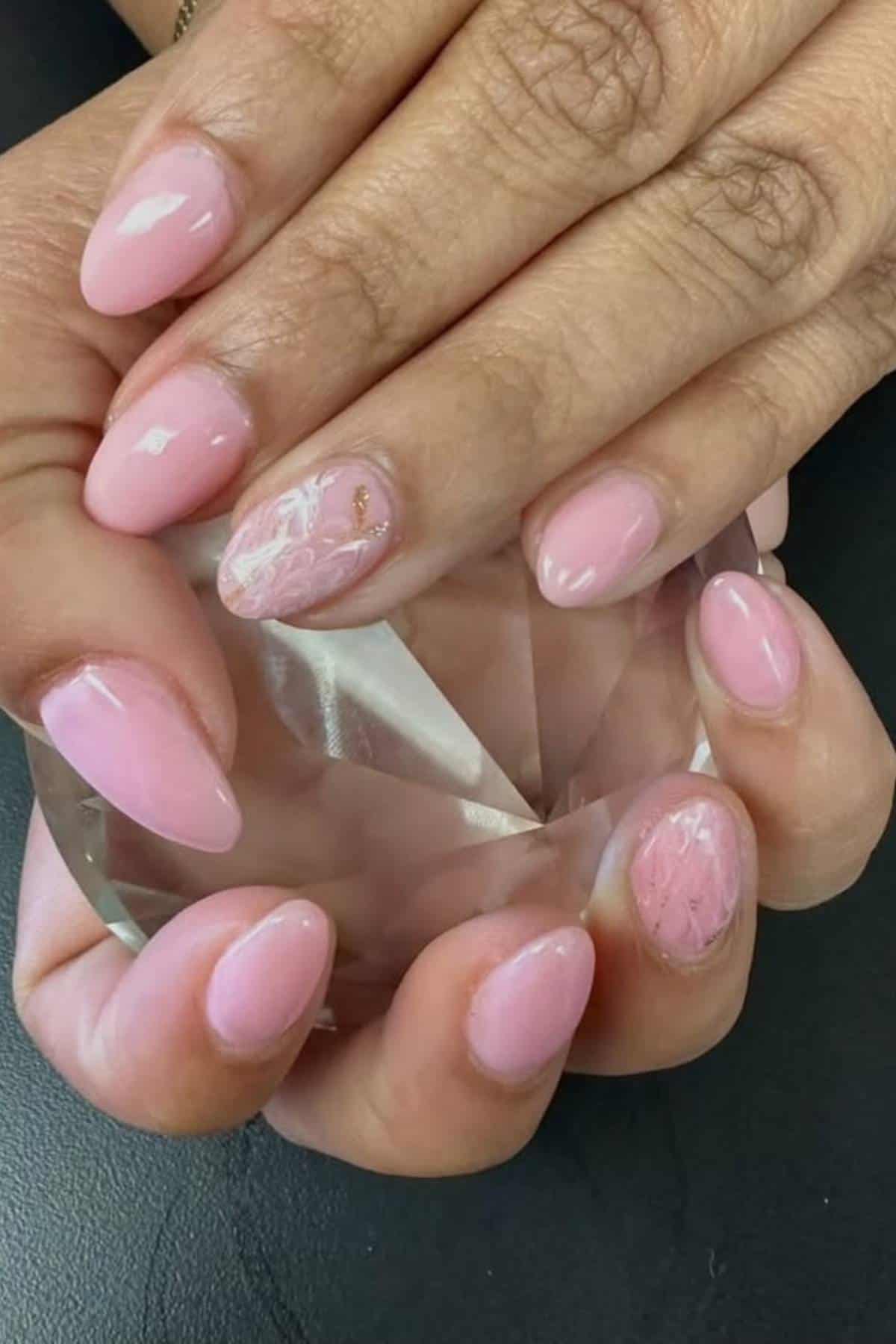 Hands with almond-shaped nails painted light pink, featuring a white floral accent design on two nails. The hands are holding a clear, faceted object.