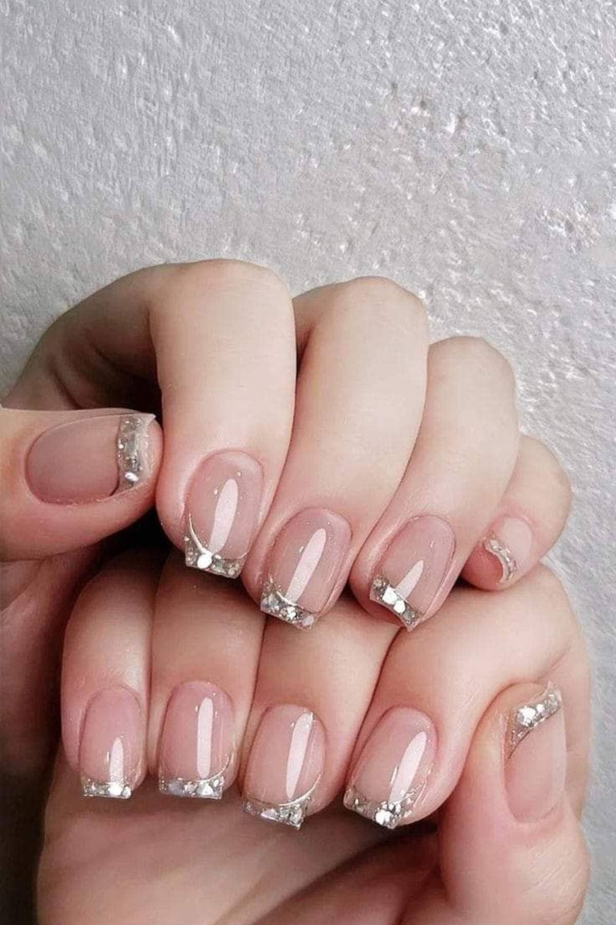 Close-up of hands with short, nude-colored nails featuring clear tips and silver glitter accents in a French manicure style.
