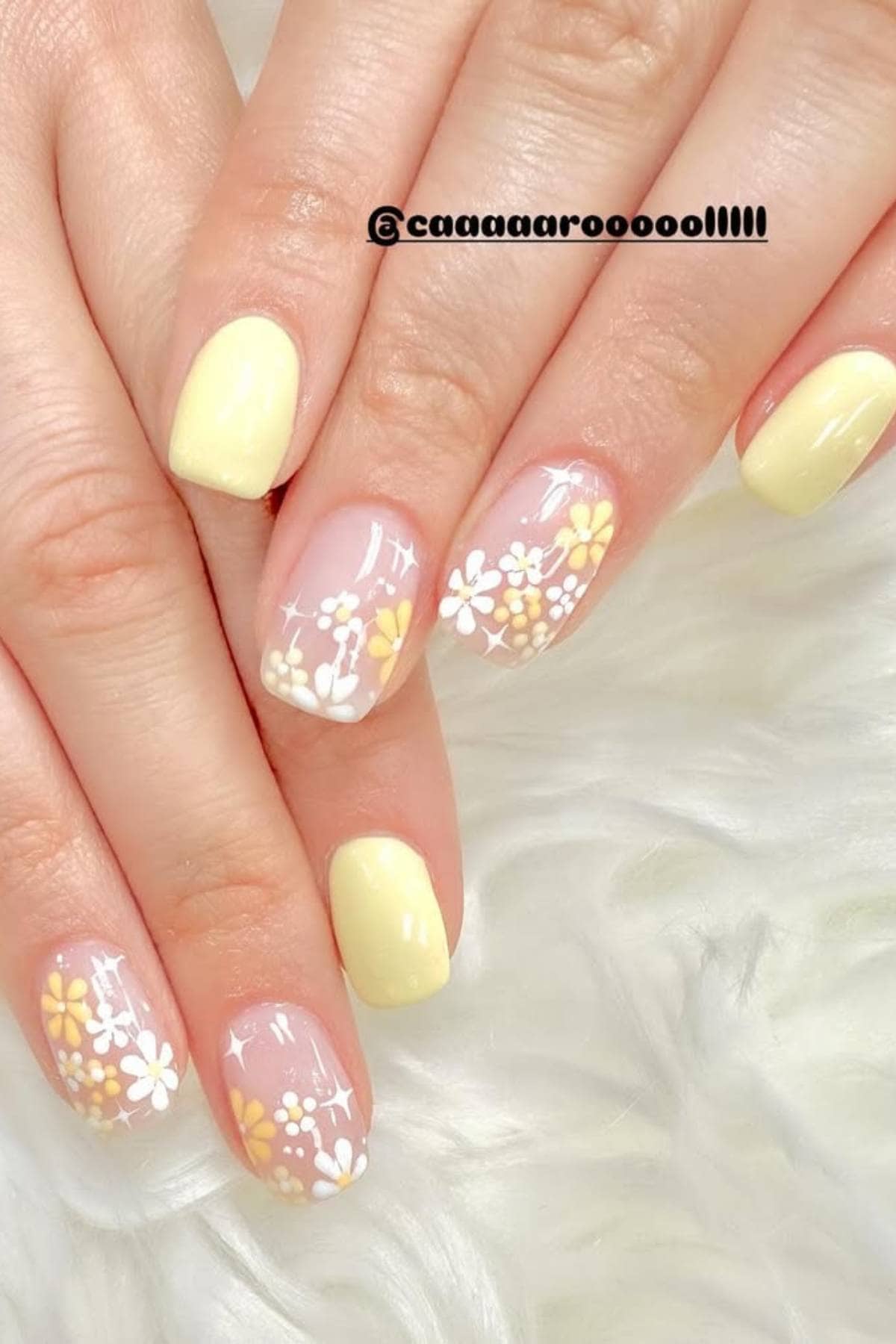 Hands with short, rounded nails painted pastel yellow and accented with clear nails featuring white and yellow daisy flower designs, resting on a white furry surface.