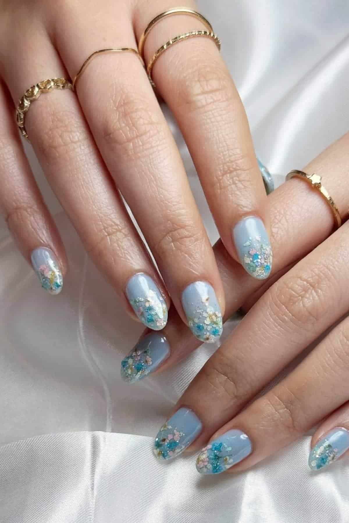 Hands with light blue almond-shaped nails featuring floral nail art and gold rings resting on a white fabric surface.